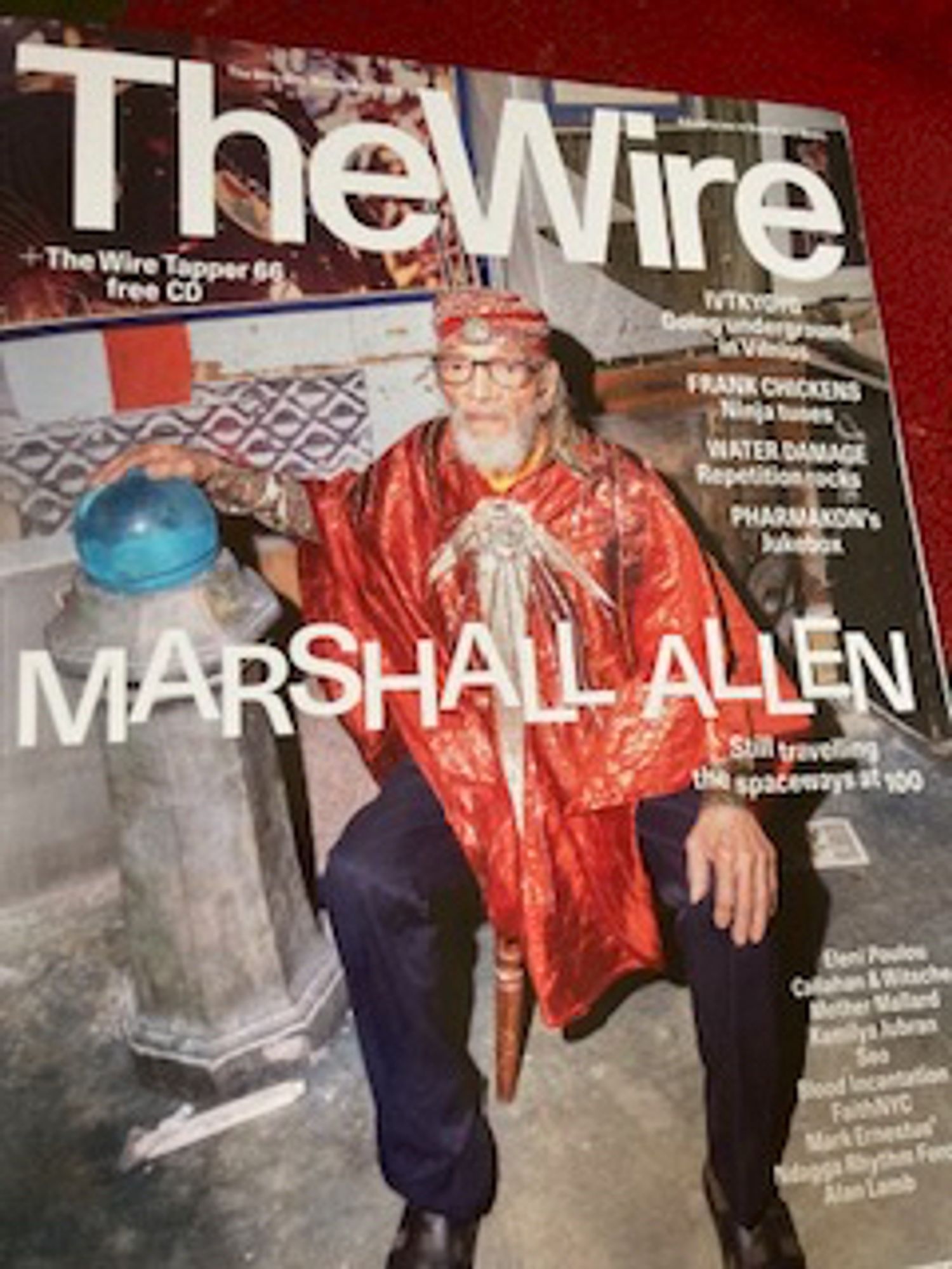 MARSHALL ALLEN on the cover of wire #489: at 100 years old, he is sat beared in an orange-&-silver robe and cap, with dark blue trousers and his hand resting on a sky-blue palantir beside him on a small stone pillar. most of the coverlines are too small to see, except for the magazine's logo and his name set in jiggled letters