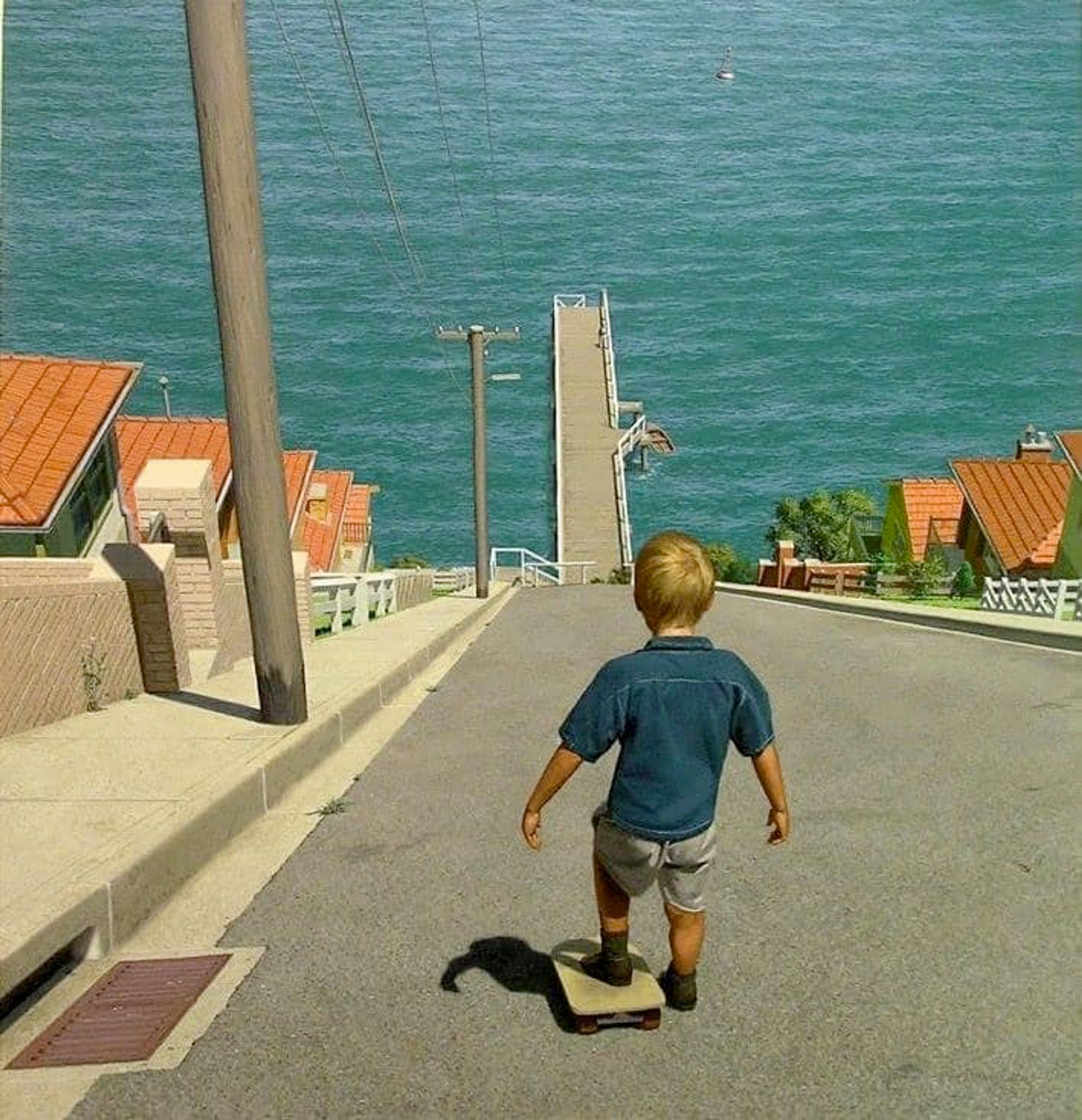 A boy stands at the top of an insanely steep hill that ends in an ocean pier. He has one foot on a skateboard.