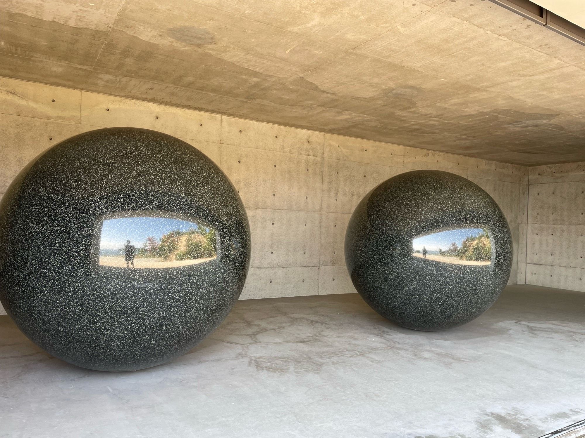 Two black marble balls about 8’ tall inside a concrete room with a side facing outside. The balls reflect the greenery of the opening and a person taking a pictute