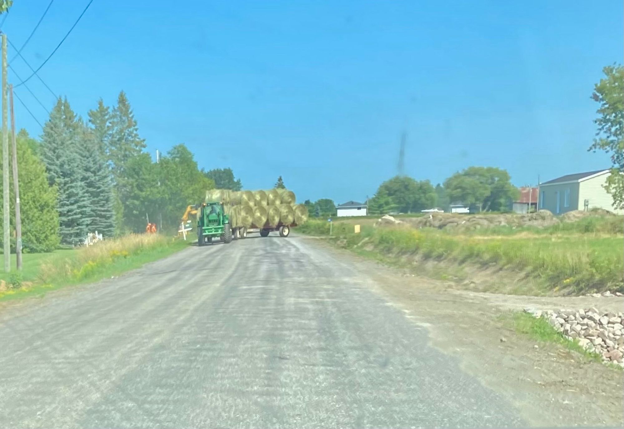 A green tractor is hauling a flatbed trailer stacked with over 10 large round bales of hay. It is leaving the farm and starting to make its journey down a gravel road