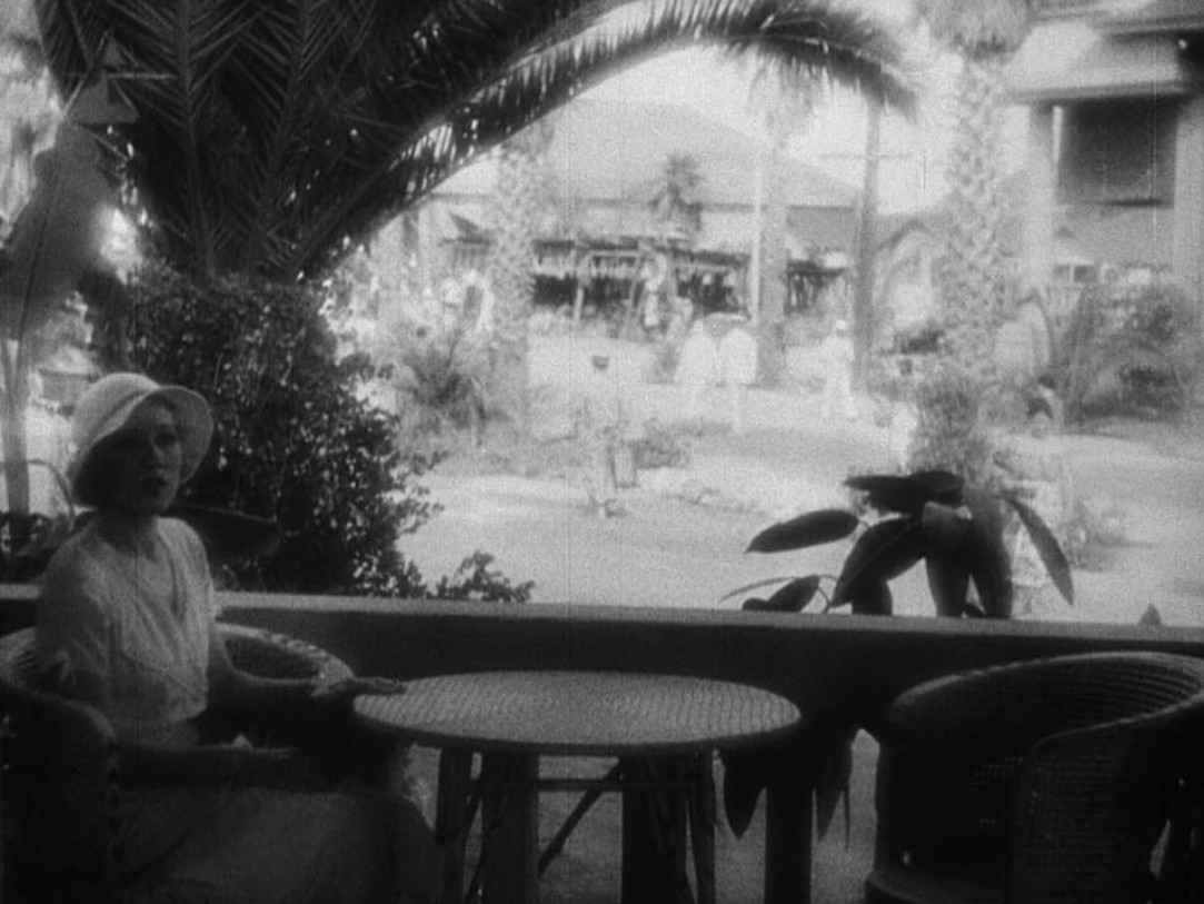 a photo of a well-dressed woman in white clothes and a hat, sitting by herself at a table, nothing on the table, buildings and tropical trees behind her