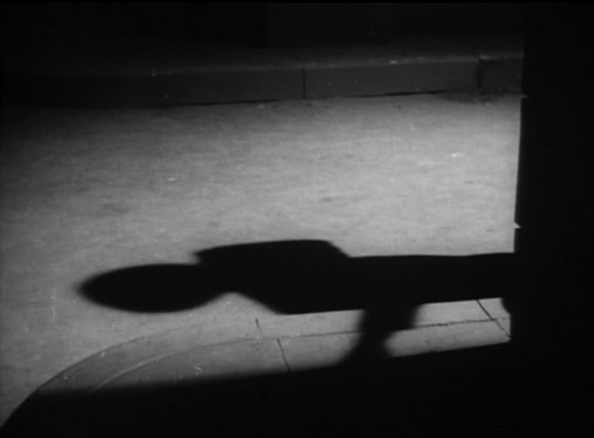 an image from the movie, a shadow is on the sidewalk and street of a person in a hat, really spooky and noir-ish