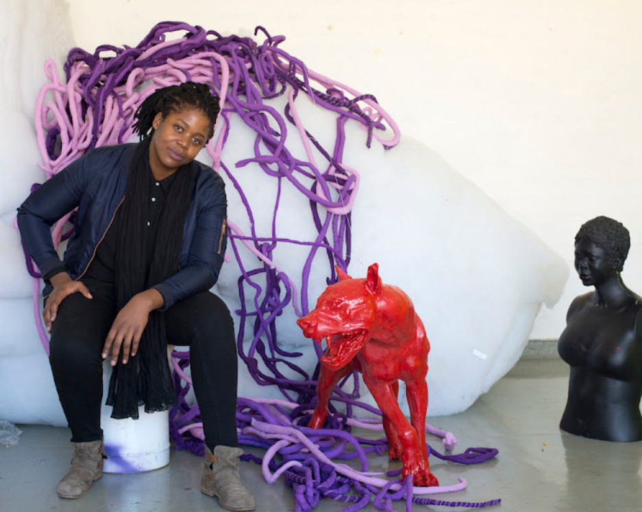 a photo of the artist, she is sitting on an upside-down bucket, a red dog statue with big teeth next to her, she is smiling warmly at the viewer, her elbow on her knee , her other hand on her leg