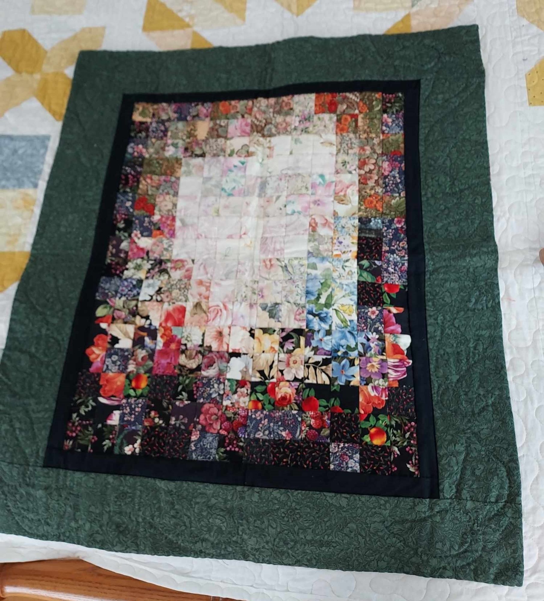 a quilt, it has a thick dark green border, a thinner black border inside, in the interior rectangle plane are floral fabrics, the lightest ones in the center, darker ones at the outer edges, creating a glowing effect 