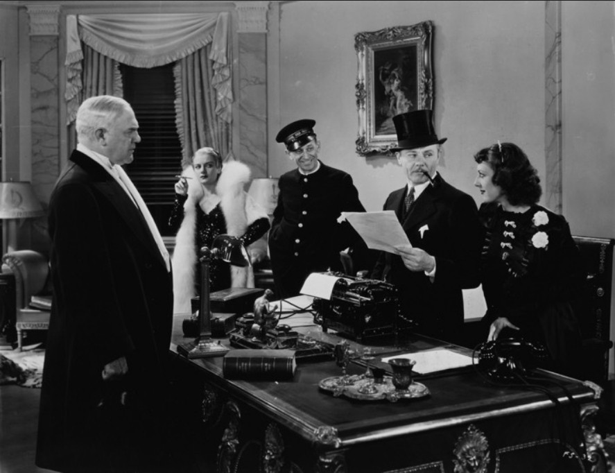 an image from the movie, the two men and their ladies, all of them nicely dressed, with the bellhop, reading papers in a finely furnished office