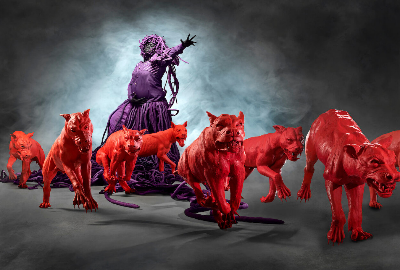 an installation, a figure in the background wearing a purple dress with snake-like pieces on her head and shoulders, the figure's arm up, commanding the red dogs in front of her to run forward, to the right of the viewer, the dogs sharp teeth, the background is grey and black tones, fog in the background