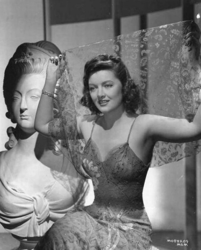 a photo of the actress, seen wearing a light dress, holding a sheer piece of fabric behind her with both hands, a bust sculpture behind her, the photo in black and white, she is looking to the left of the viewer, seen in a three quarter view, a faint smile on her face