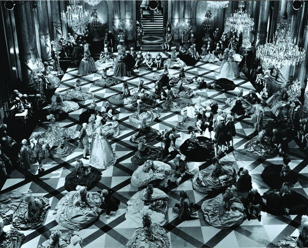 an image from the movie, an aerial view of a ballroom dance, women's large hoop dresses twirling around on the dance floor, men dancing with them in darker clothing , the pattern of the tile floor is diagonal black lines over squares, chandeliers are hanging down