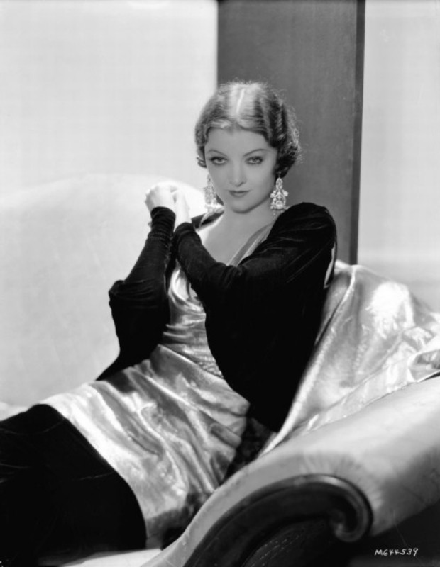 a photo of an actress, black and white, taken in the 1930s, she is wearing a dark sweater and skirt with gold dress over it, she has short wavey flapper-style hair, a white background with dark rectangle behind her, she is sitting on a large comfortable chair, her hands together at her collarbone, looking alluringly at the viewer