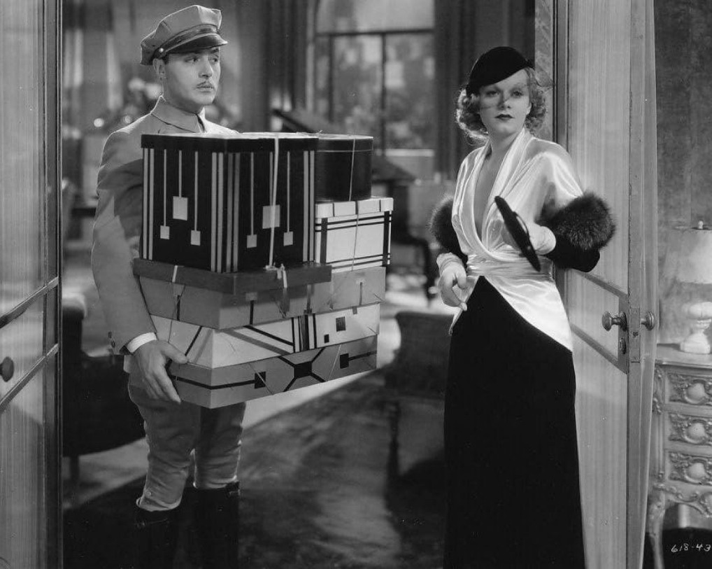 an image from the movie, a doorman is carrying a large box , he is standing at the entrance to a fashionably dressed woman's apartment, she is looking around the room, deciding where to tell him to put it