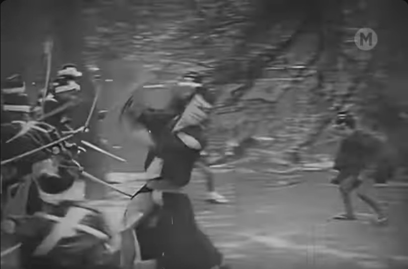 an image from the movie, it is in black and white, a figure in a black robe is whirling with a sword, fighting off attackers, some trees behind them, a figure in dark clothing in the background