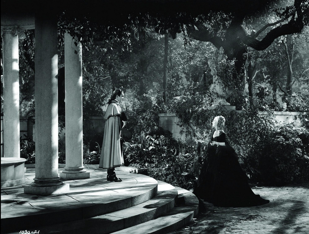 an image from the movie, it is night time, a man is standing on the front steps of his palace, large columns behind him, he is seen from the side, wearing a long cloak, the moonlight shining through, a woman in a black dress is talking to him, facing each other , there are trees behind them and shrubs