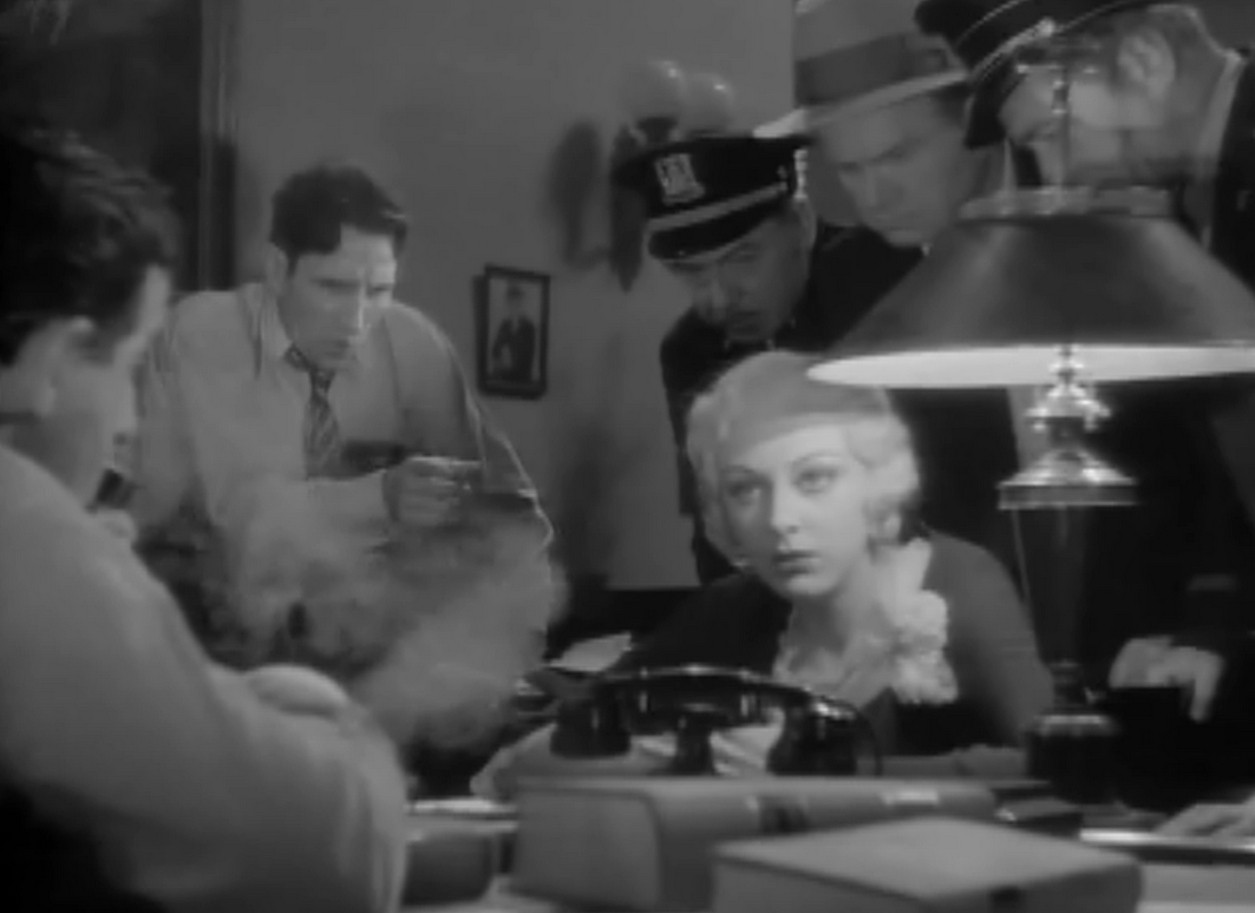 a woman talking over a desk to a man, theyre both seated, other men seated around them in the office, several police officers and detectives