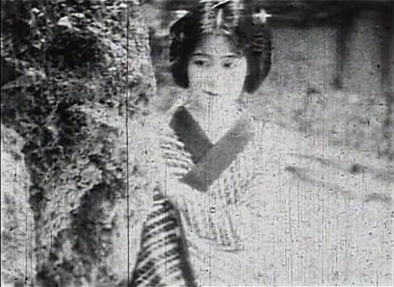 a woman in a kimono looking out from behind a  tree, her hand on it