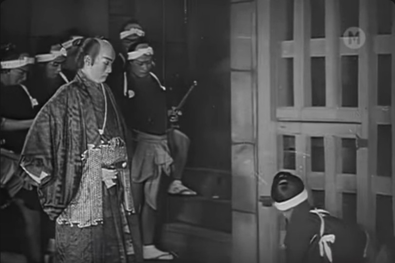 an image from the movie, a man in elaborate robes standing in front of the wooden doors of a jail cell, there are other warriors behind and around him, looking inside the prison cell