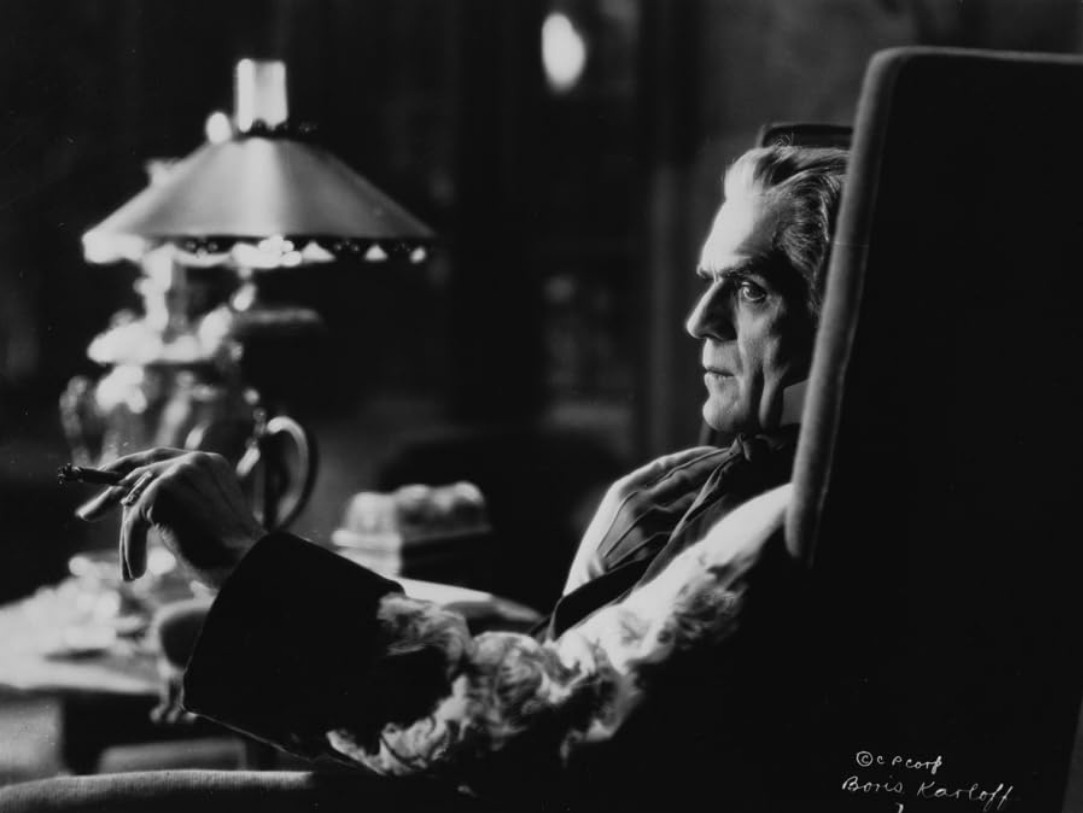 boris karloff as the main character, seen from the side sitting in a chair, a cigar in his hand, an alert expression on his face, a desk with a lamp on it behind him 