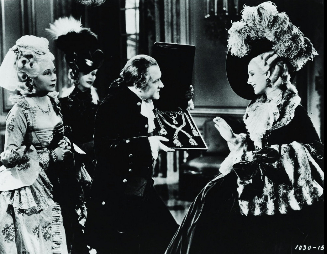 an image from the movie, a short stout man in fancy clothing presenting a fine set of large jewels to a woman wearing a large hat with feathers, the women standing behind the man are dressed extravagantly, the woman getting the jewelery is smiling , she is seen from the side, wearing a large hoop dress with lots of ruffles and layers on it, she wears gloves on her hands