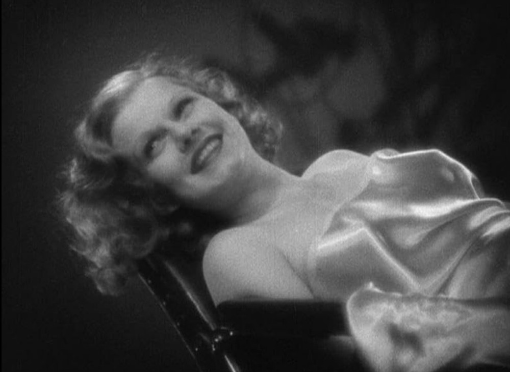 an image of the star of the film, in a scene, reclined in a sofa chair, smiling, looking to her upper right, wearing a silk / satin dress