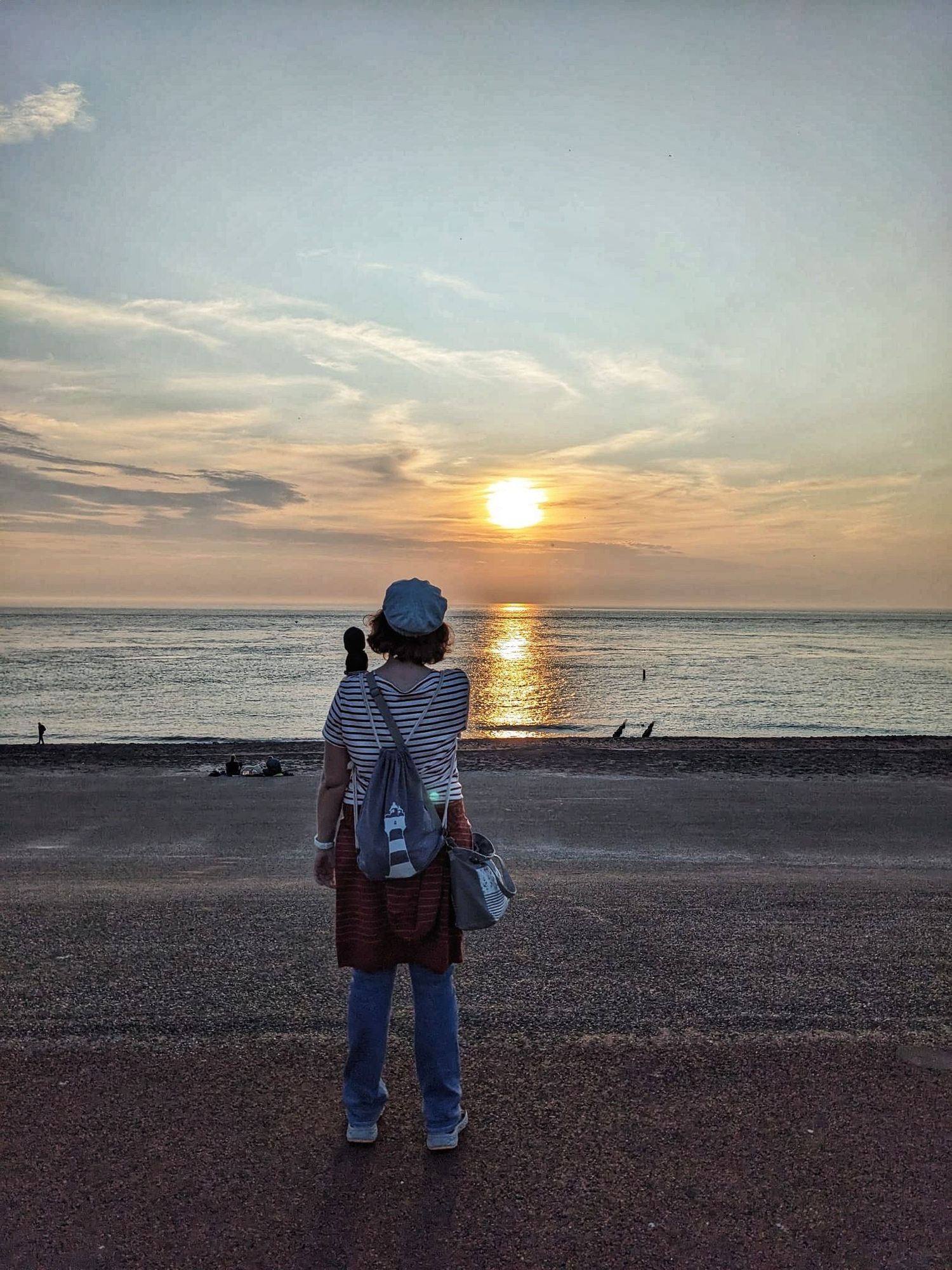 Ein Foto von mir vor dem Sonnenuntergang über der Nordsee. Man sieht mich von hinten. Ich trage ein gestreiftes Shirt, Jeans und ein Käppi. Auf dem Rücken habe ich einen Turnbeutel mit Leuchtturm und ein Plüschtier sitzt auf meiner Schulter. Der Sonnenuntergang ist blau-gelb-orange.