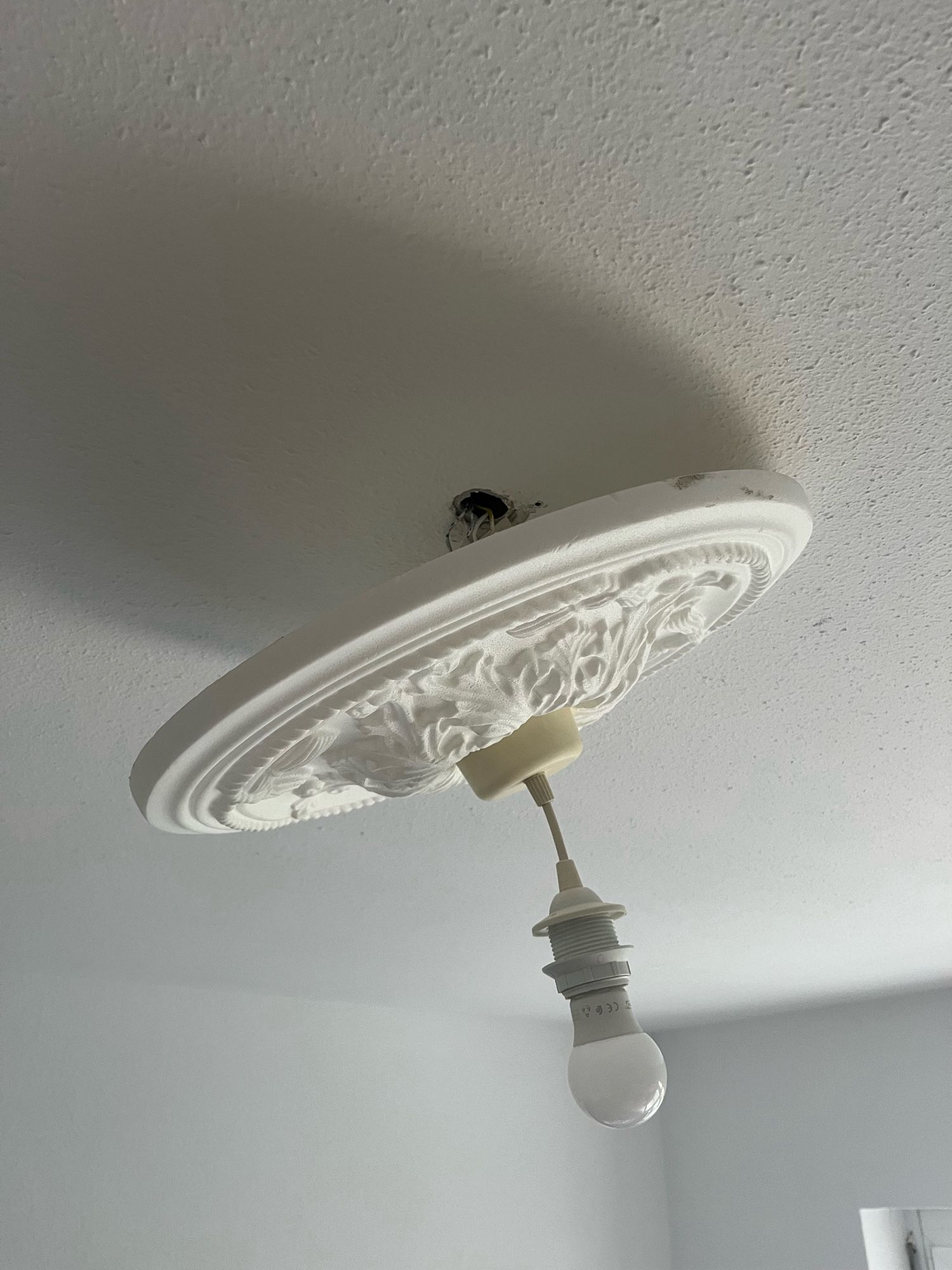 Man sieht eine, sehr schief von der Decke hängende Lampe samt Leuchte, die nicht so halten wollte, wie geplant. Schief hängt sie, weil ein Kreisrundes Decken-Styropor mit Stuck Optik sich verkanntet hat.