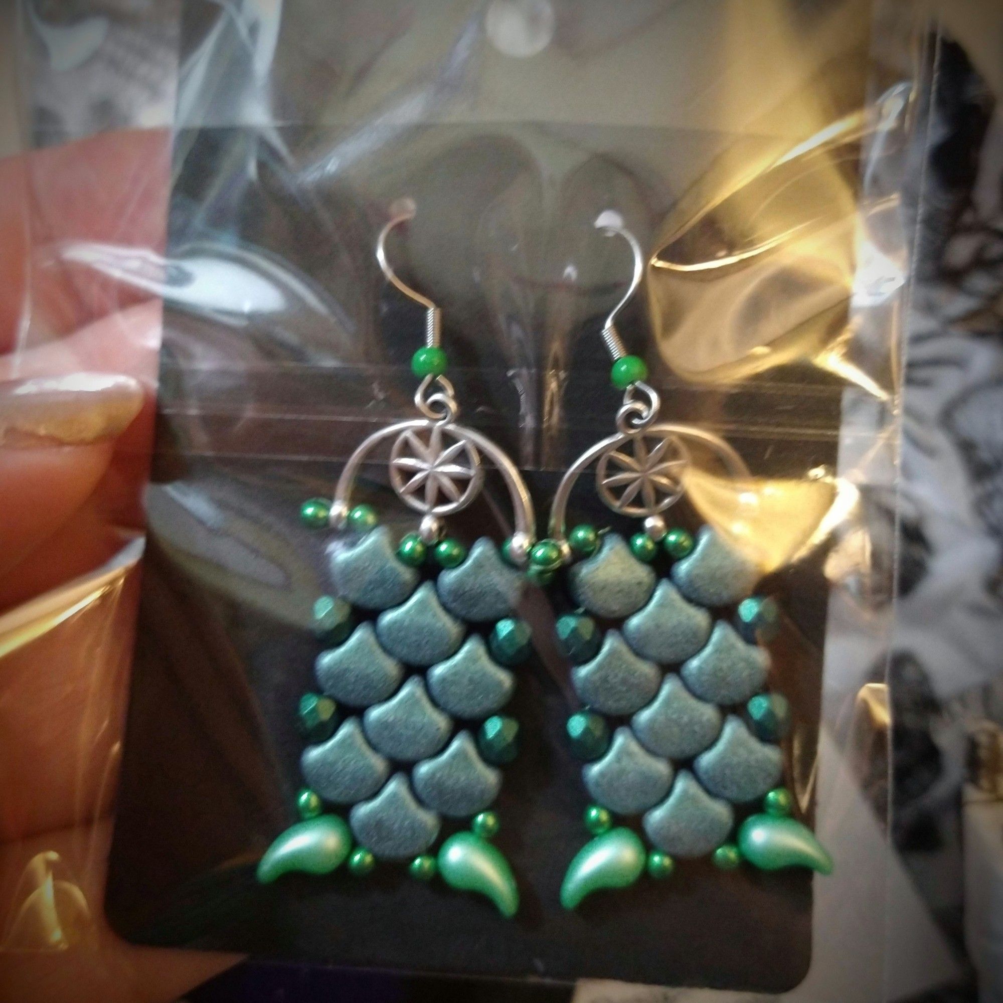 A pair of dangly green and silver earrings in a plastic envelope. The main body of the earring design is made of muted metallic green beads shaped like flat drops; they're sewn together in a tessellated pattern that looks like fish or dragon scales, and finished at the bottom with a different type of pearlescent green bead that looks like claws coming out of the bottom corners. The beadwork part is suspended from an arched silver frame with what looks a bit like a stylized compass rose in the center of the arch, and the whole thing hangs from silver fish-hook wires with little green accent beads.