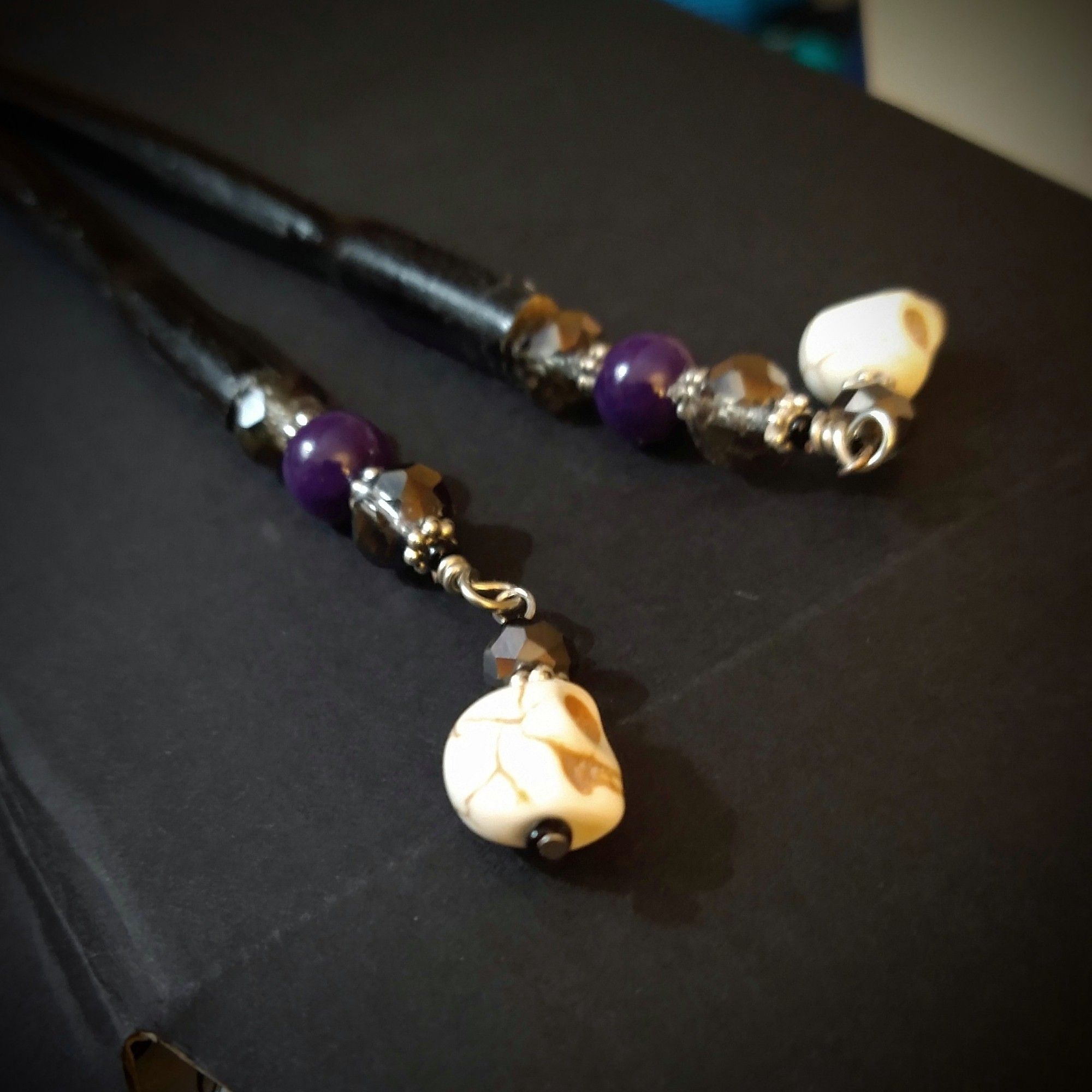 A pair of black wooden hair sticks topped with smoky grey faceted glass beads, deep purple-dyed jade spheres, and deliciously dangly skull charms made of ivory-colored howlite.