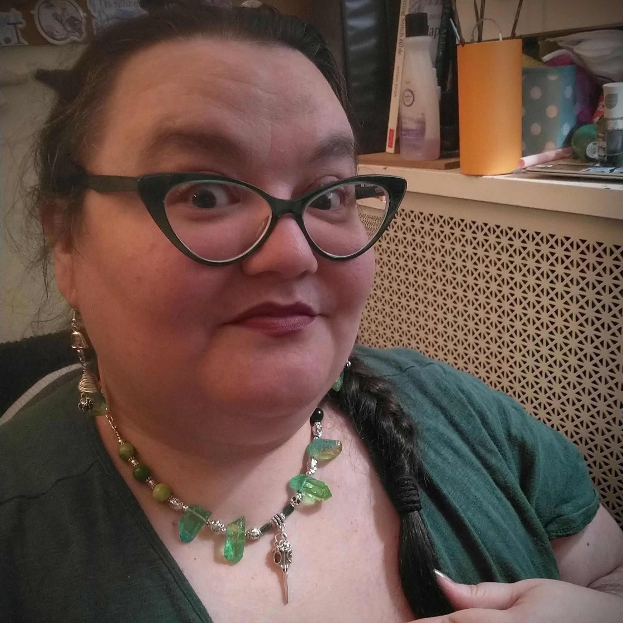 A fat Arab-American brunette in a dark green dress and matching glasses, looking super-stoked to be wearing a matched green-and-silver earring-and-necklace set. The necklace is a bit longer than a choker, made of big jagged chunks of green-dyed quartz alternating with silver skull spacer beads and culminating in a raven skull pendant. The large, dangly earrings aren't super visible in this spot, but it's clear they're made of the same green quartz chunks and silver beads and findings.