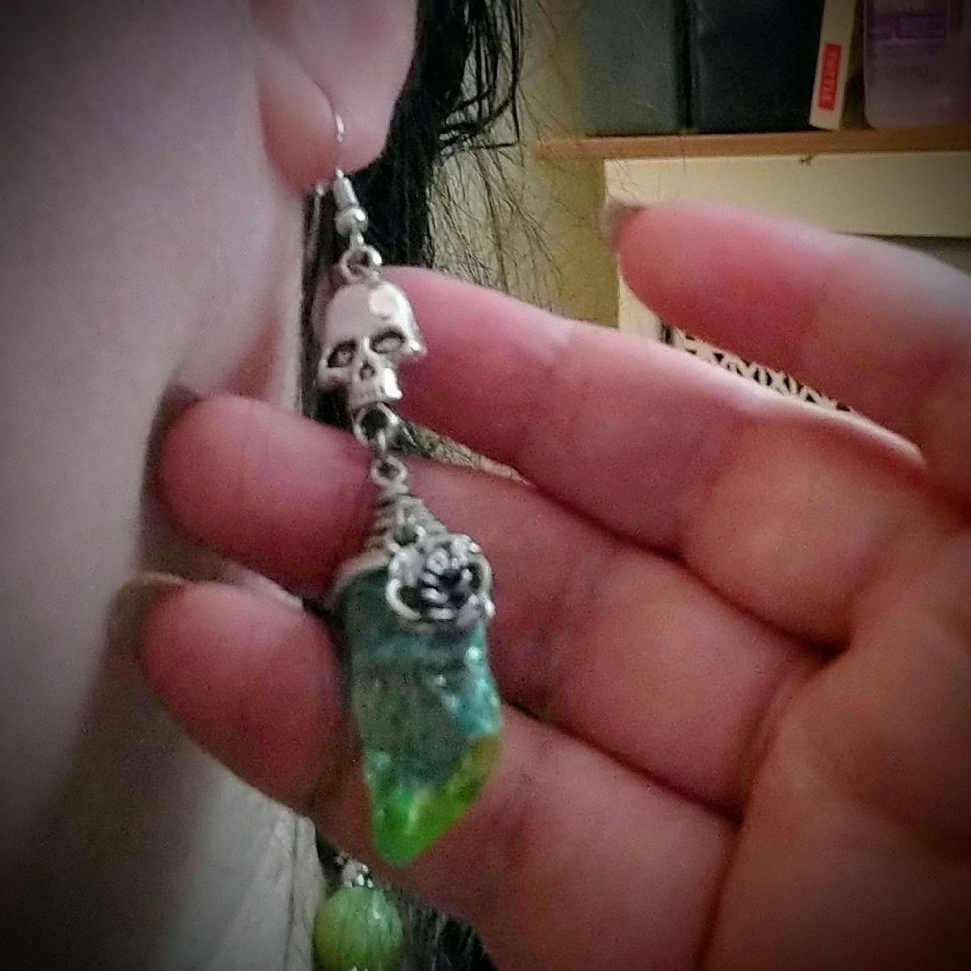 A close-up of the big green quartz earrings. The stone chunk is wrapped with silver wire around the top and features a silver rose charm dangling in front. The assembly is suspended from a big silver skull charm.