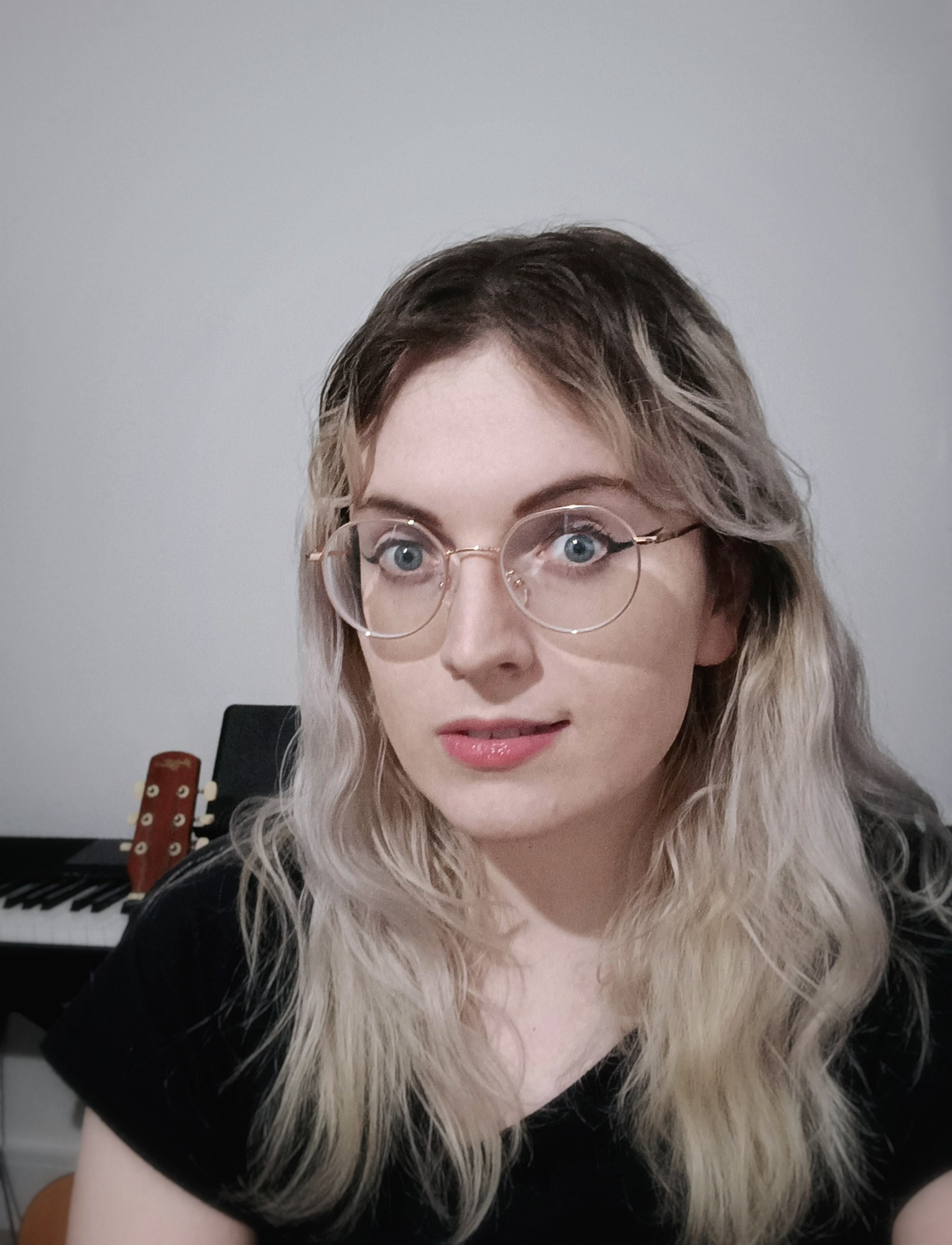 me sat in front of a piano and a guitar, I am slightly smiling. I am wearing round frame glasses and eyeliner