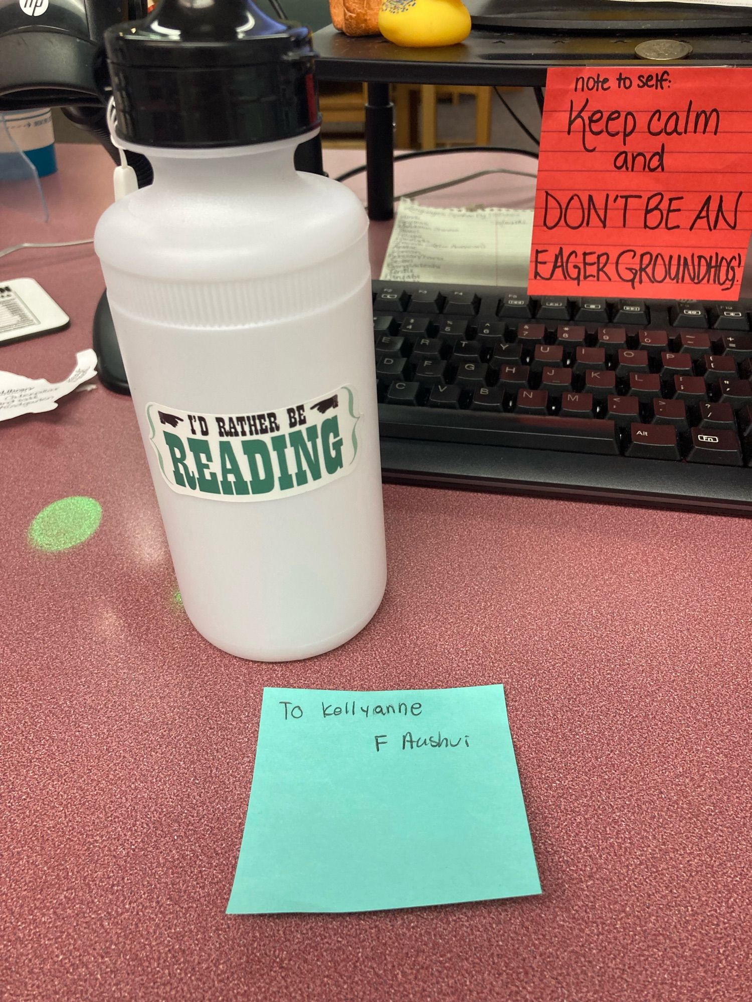 A white water bottle with the sticker “I’d Rather Be Reading” sits on my work desk with a light aqua colored post-it note that says “To Kellyanne, F Aashvi”