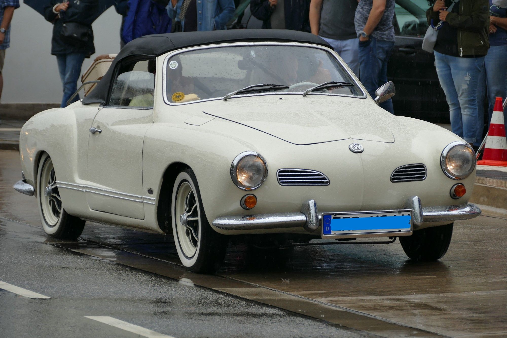 Das Bild zeigt ein vintage weißes Cabriolet mit einem schwarzen Stoffverdeck, das auf einer nassen Straße geparkt ist. Das Fahrzeug hat markante runde Scheinwerfer, verchromte Stoßstangen und ein blaues Nummernschild vorne. Im Hintergrund sind Menschen zu sehen, was darauf hindeutet, dass dies bei einer Veranstaltung oder einem öffentlichen Treffen aufgenommen wurde. Das Design des Autos deutet auf einen Stil aus der Mitte des 20. Jahrhunderts hin.