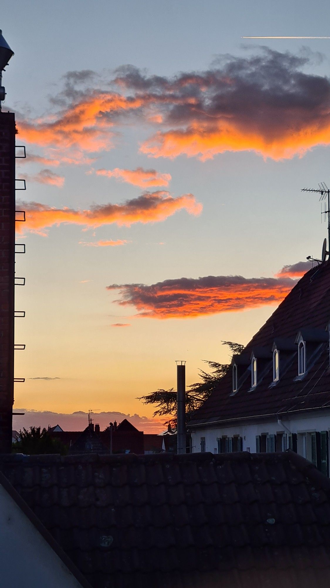 Das Bild zeigt einen Sonnenuntergang mit einem lebhaften orange-blauen Himmel, der zwischen zwei silhouettierten Strukturen, möglicherweise Gebäuden, zu sehen ist. Die Wolken sind vom untergehenden Sonnenlicht erleuchtet, was eine dramatische und farbenfrohe Szene erzeugt.