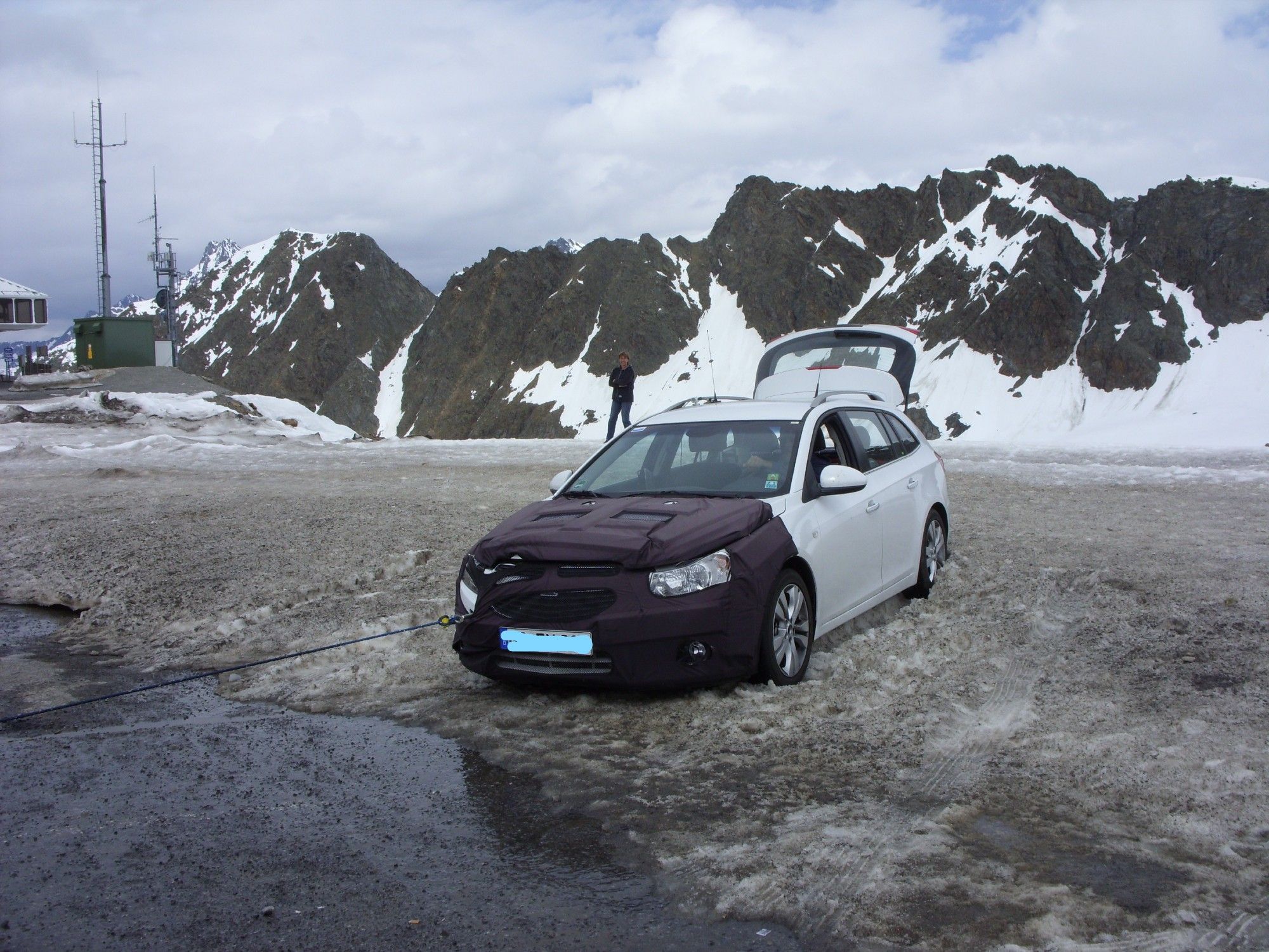 Das Bild zeigt ein dunkles Auto mit geöffnetem Motorhaube, das mit einem Abschleppseil an ein anderes Fahrzeug angeschlossen ist. Dies deutet darauf hin, dass das Auto eine Panne hat oder feststeckt. Der Boden ist nass und scheint eine Mischung aus Schnee und Wasser zu sein, was auf eine kalte oder bergige Gegend hinweist. Im Hintergrund sind schneebedeckte Berge und ein klarer Himmel mit einigen Wolken zu sehen. Auf der linken Seite des Bildes befindet sich eine Struktur, die wie eine Antenne oder ein Kommunikationsturm aussieht. Eine Person geht von der Kamera weg in Richtung anderer Strukturen in der Ferne.