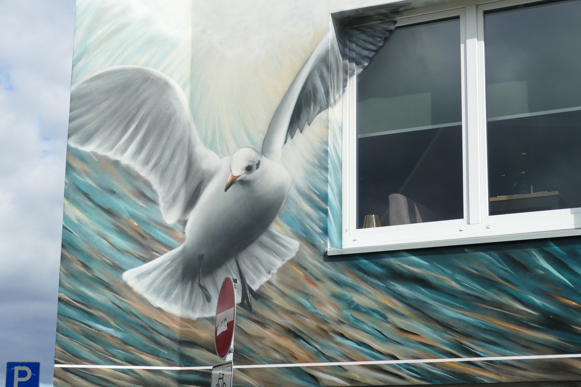 Das Bild zeigt ein großes Wandgemälde einer fliegenden Möwe an der Seite eines Gebäudes. Die Möwe ist mit weit ausgebreiteten Flügeln dargestellt und wirkt sehr lebensecht. Der Hintergrund des Wandgemäldes hat Blautöne und Weiß, die Wellen im Ozean ähneln. Ein echtes Fenster ist in das Design integriert und passt zu der Stelle, an der der Flügel des Vogels angehoben ist, was eine interessante Interaktion zwischen dem Kunstwerk und dem Gebäude schafft. Vor diesem Wandgemälde befindet sich ein Parkplatzschild mit einem “P”-Symbol, das auf Parkmöglichkeiten hinweist, und einem Pfeil, der nach unten zeigt. Unter diesem Schild ist ein weiteres Schild teilweise sichtbar, das rot-weiße Farben hat und möglicherweise eine Art Einschränkung oder Warnung im Zusammenhang mit dem Parken anzeigt.