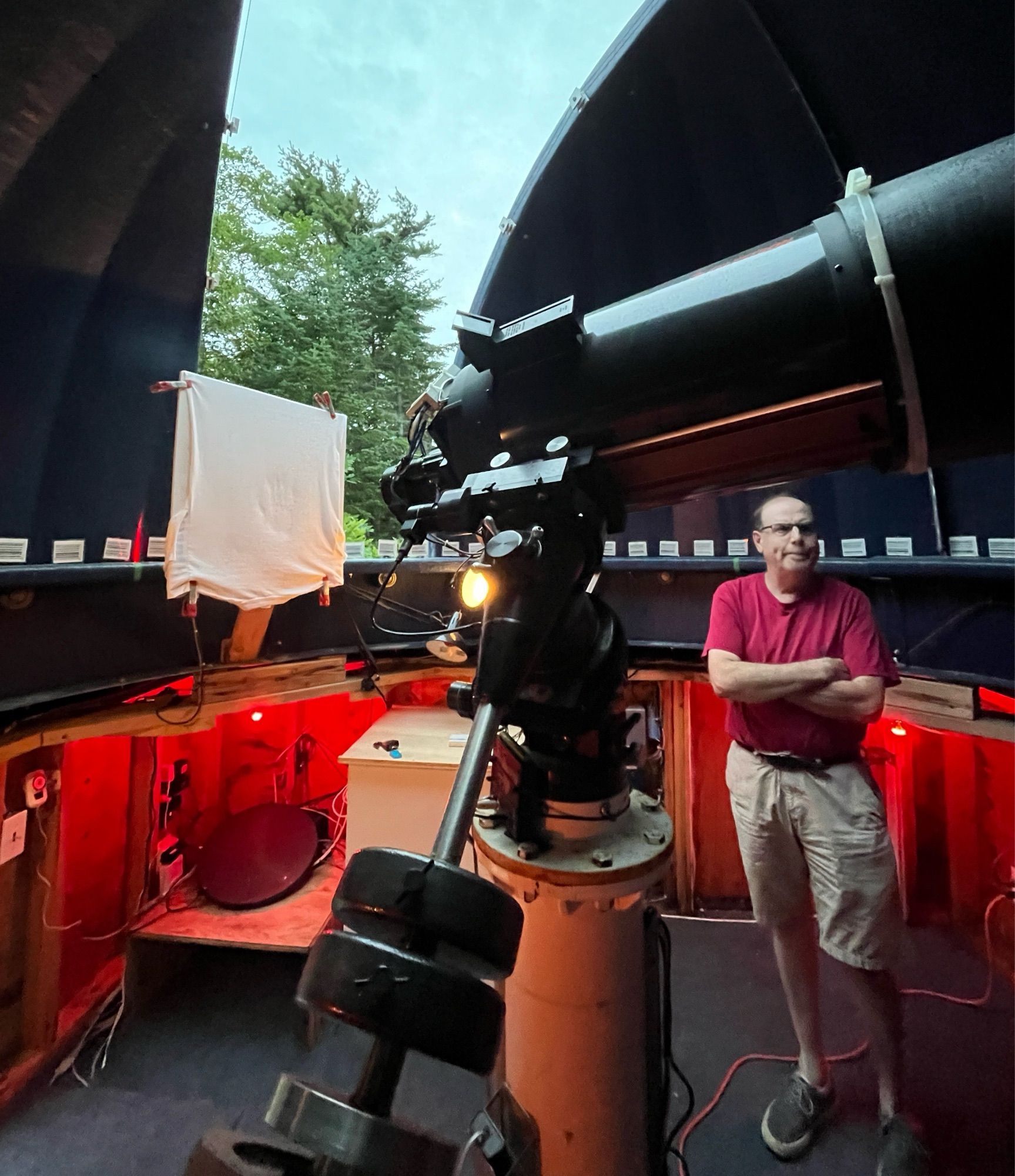 Image of Dave Lane standing inside of his backyard observatory, the Abbey Ridge Observatory. The inside of the observatory dome has dark ceilings and there are red lights lighting up the lower half of the dome inside. There is a telescope on a pier in the middle, in front of Dave.