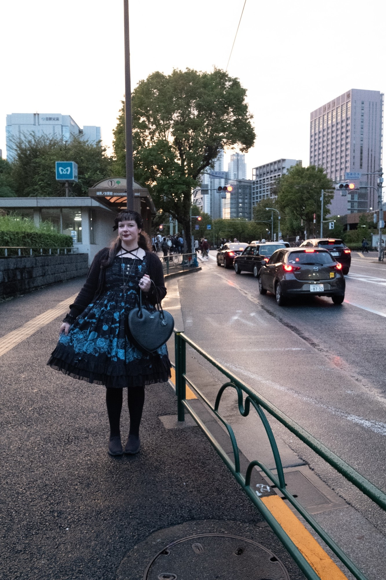 Woman (me) standing on the footpath alongside a road with cars. Posing wearing a black and blue outfit.