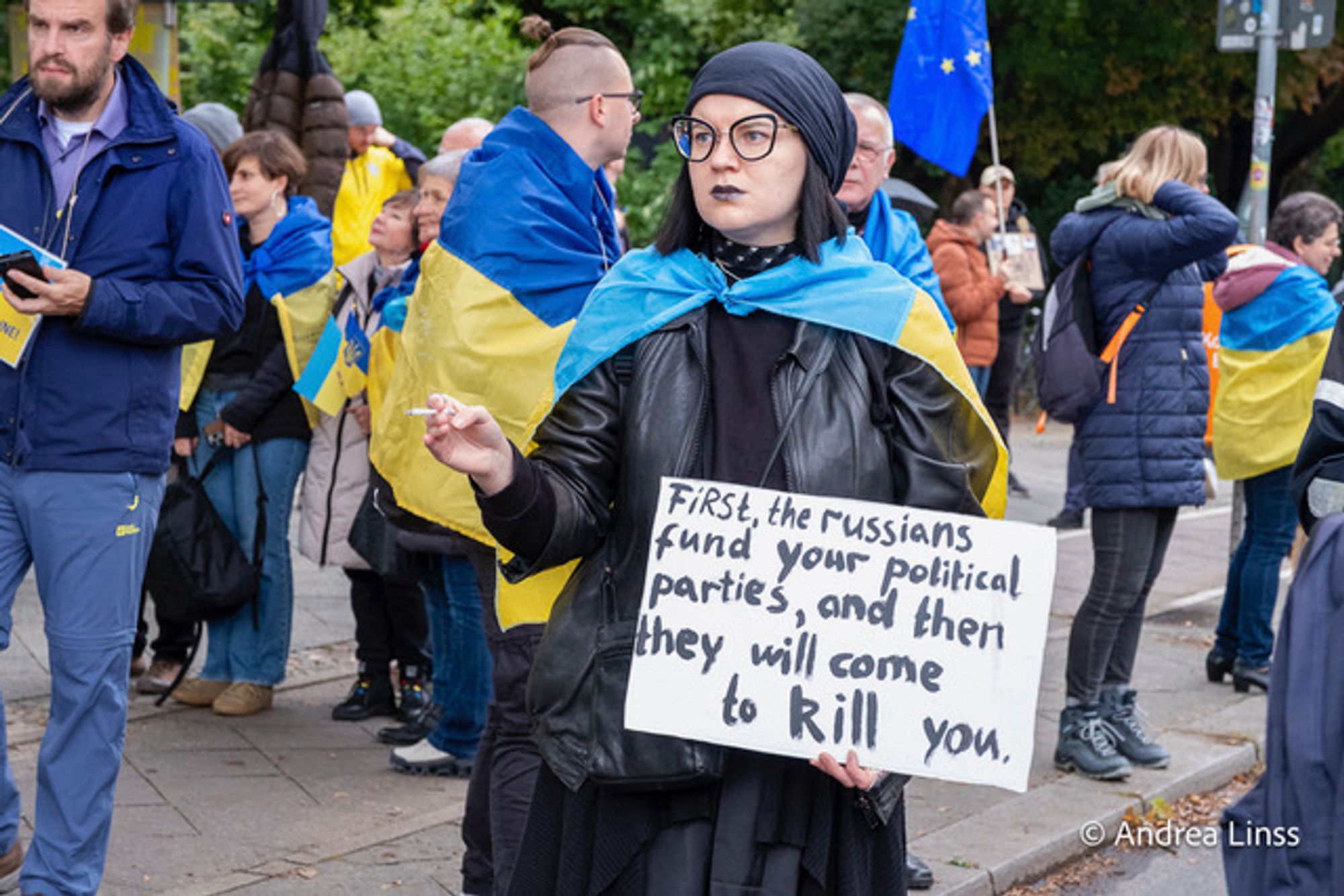 Eine junge Frau in schwarz, mit schwarzer Brille , in einer Hand eine Zigarette, in der anderen ein Plakat, auf dem „ First the russians fund your political parties and then they will come to kill you „ zu lesen ist.