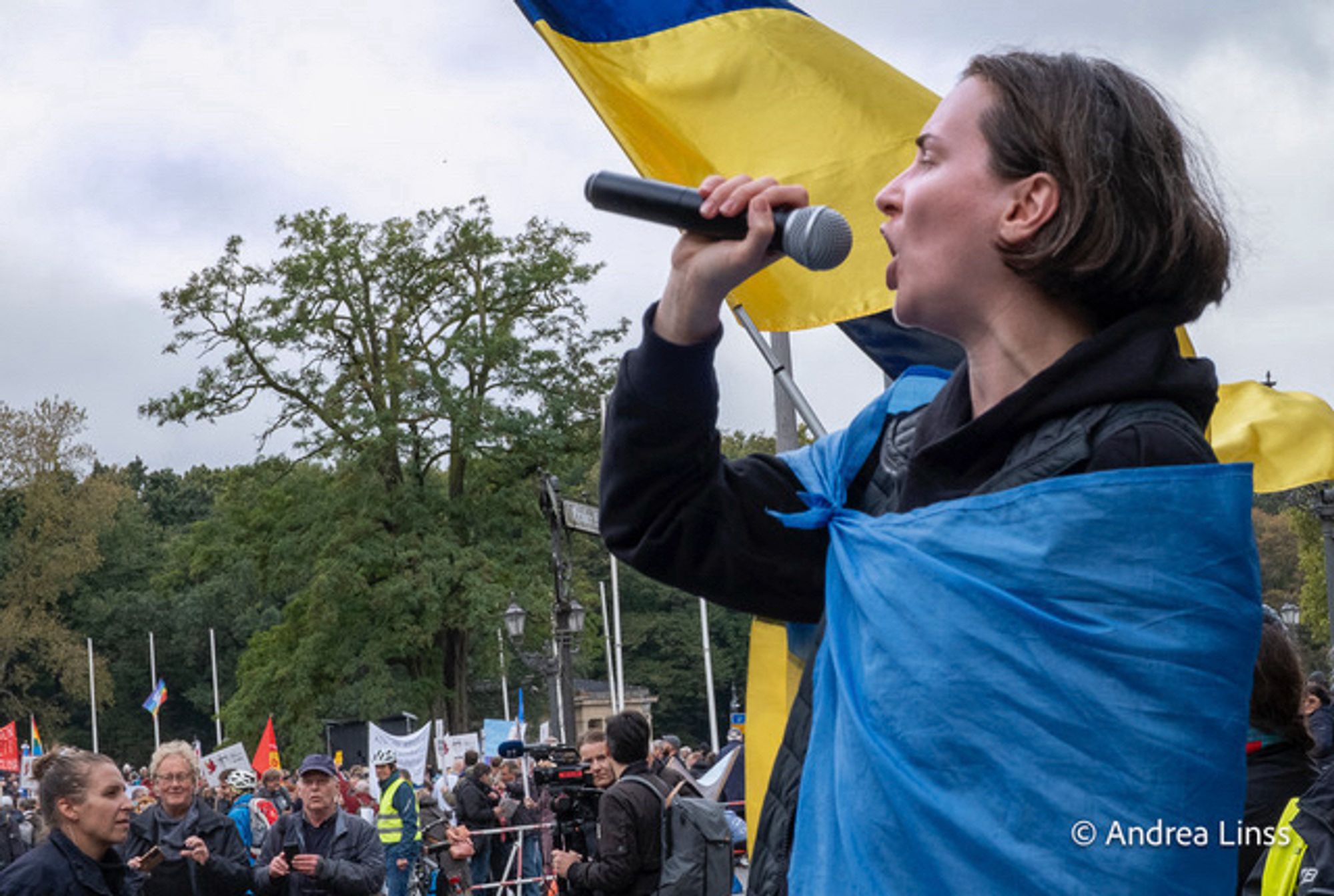 Eine junge Frau ruft in ein Mikrophon, sie hat eine ukrainische Fahne ist blau-gelb umgehängt, sie steht vor einer ukrainischen Fahne.