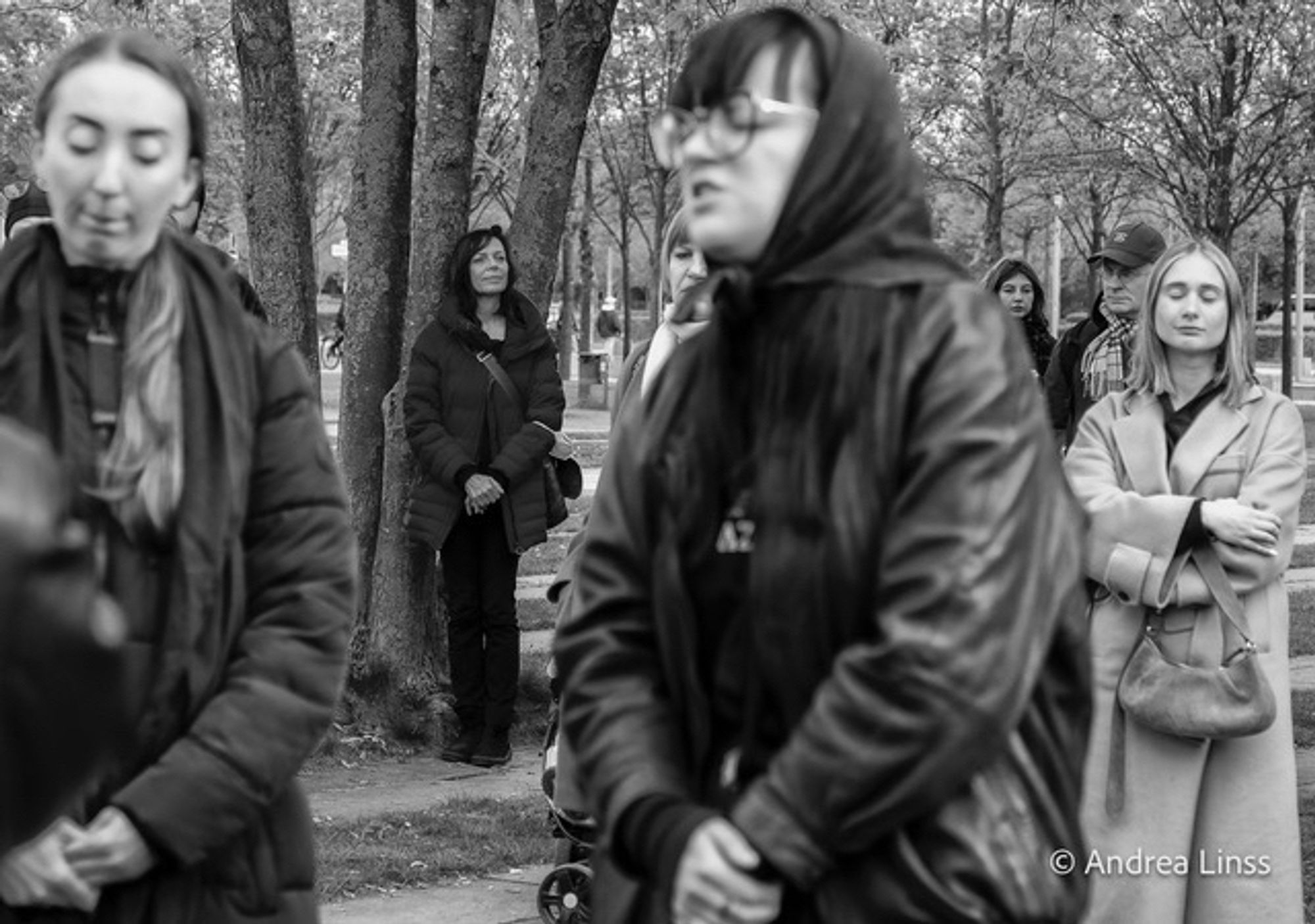 Schwarzweißfoto. Im Vordergrund, unscharf, zwei schwarz gekleidete junge Frauen. Hinter ihnen, an einen Baum gelehnt eine ebenfalls schwarz gekleidete Frau, sie schaut gedankenverloren vor sich hin. Rechts im Hintergrund eine junge blonde Frau, mit hellem Mantel, die Arme vor sich verschränkt, eine Handtasche haltend , sie hat ihre Augen verschlossen.