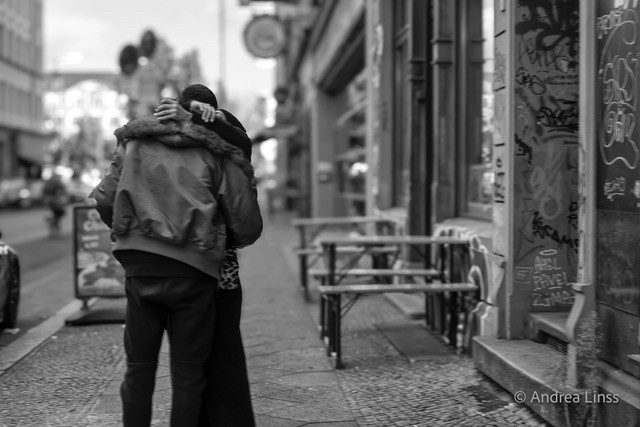 Schwarzweißfoto. Straßenszene. Ein Mann und eine Frau umarmen sich . Rückenansicht vom Mann , die Frau wird von ihm verdeckt. Rechts Altbauten , ein Hauseingang mit Graffitis, im Hintergrund Biertische und Bänke.