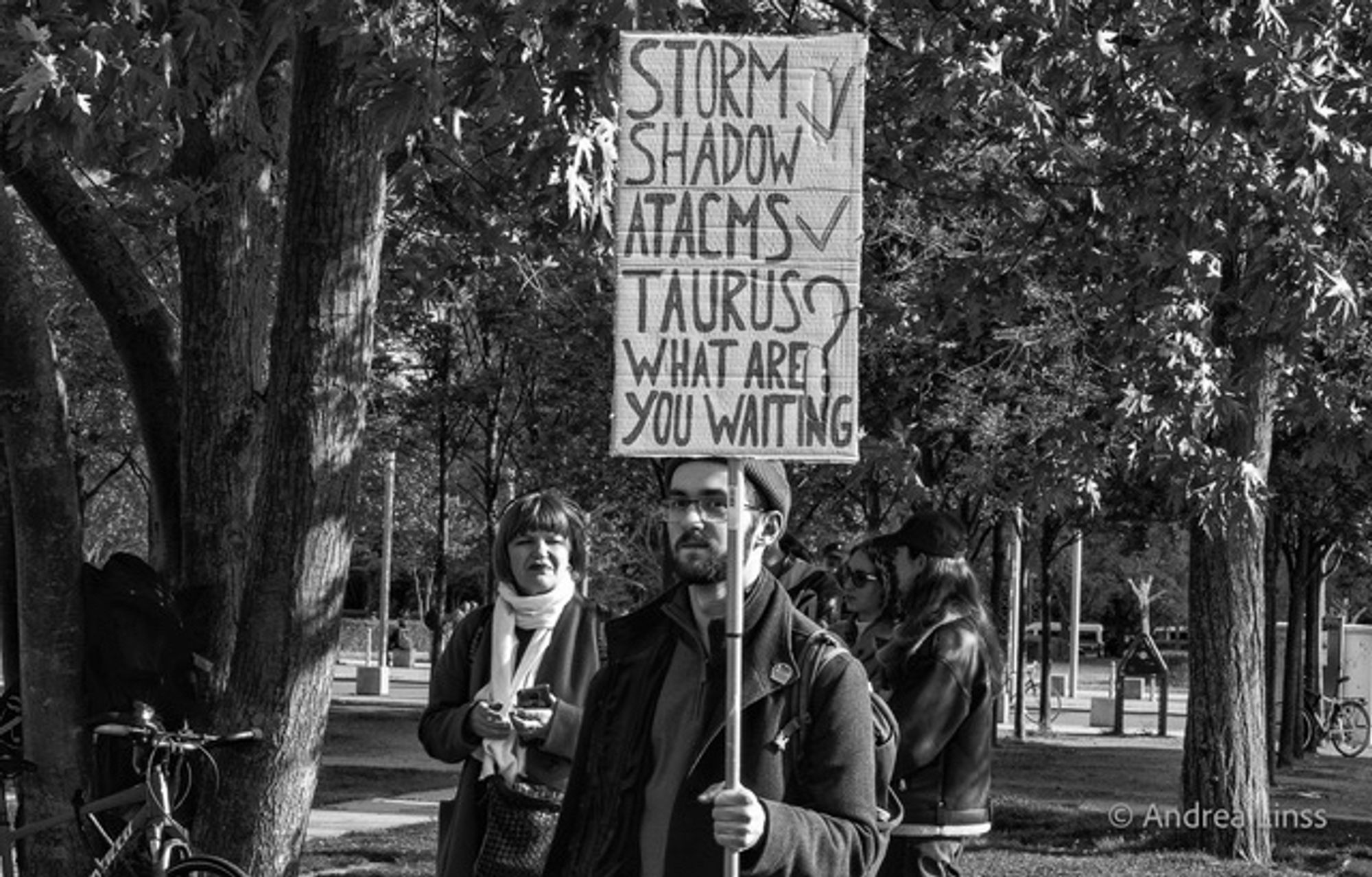 Schwarzweißfoto. Viele Bäume, Im Vordergrund, mittig, ein junger Mann , mit Brille, Wollmütze, Bart, dunkle Jacke. Er schaut zur Kamera, in der linken Hand hält er an einem langen Stab ein Schild, auf dem „Storm Shadow mit Häkchen, Actams mit Häkchen, Taurus ? What are you waiting for „ zu lesen ist. Hinter ihm zwei Frauen, eine schaut nach hinten, die Andere Richtung Kamera.