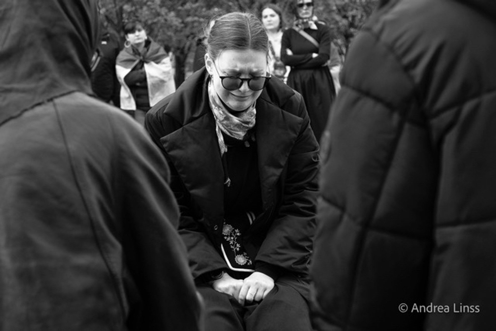 Schwarzweißfoto. Eine junge schwarz gekleidete Frau, auf dem Boden kniend, beide Hände vorne auf ihren Beinen, sie trägt eine dunkel gefärbte Brille, schaut nach unten und weint.