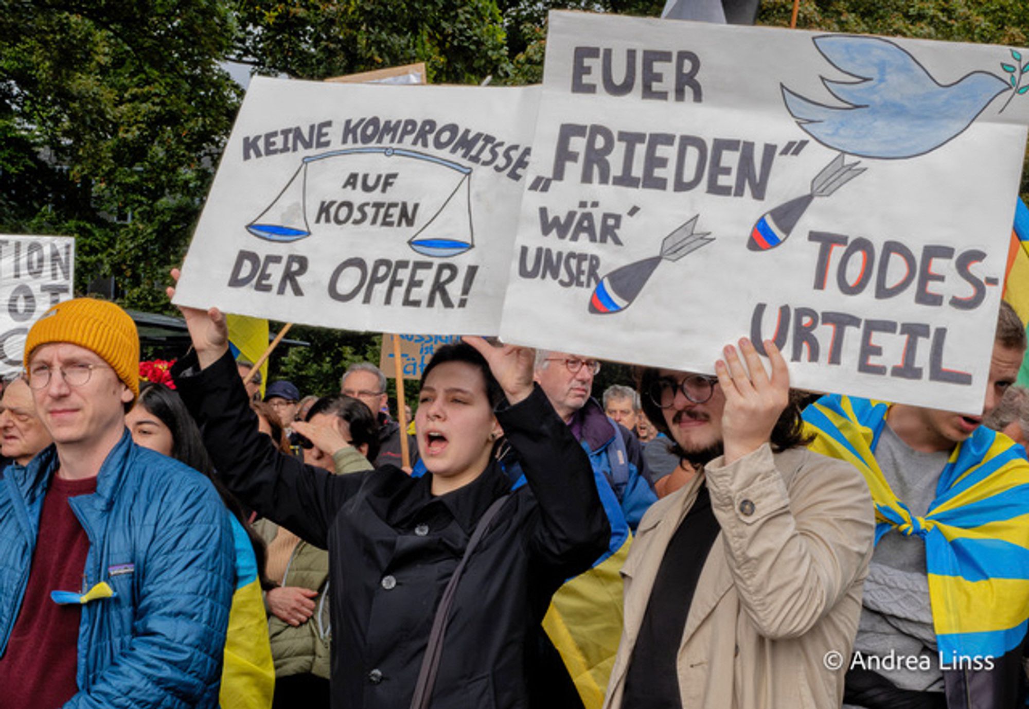 Viele Menschen. Im Vordergrund ein junger Mann und eine junge Frau mit jeweils einem Plakat , das sie hochhalten. Auf dem Plakat der Frau ist eine Waage zu sehen, mit dem Text „ Keine Kompromisse auf Kosten der Opfer „. Auf dem zweiten Plakat ist eine Friedenstaube, die Bomben abwirft, zu lesen ist „ Euer Frieden wär unser Todesurteil“.