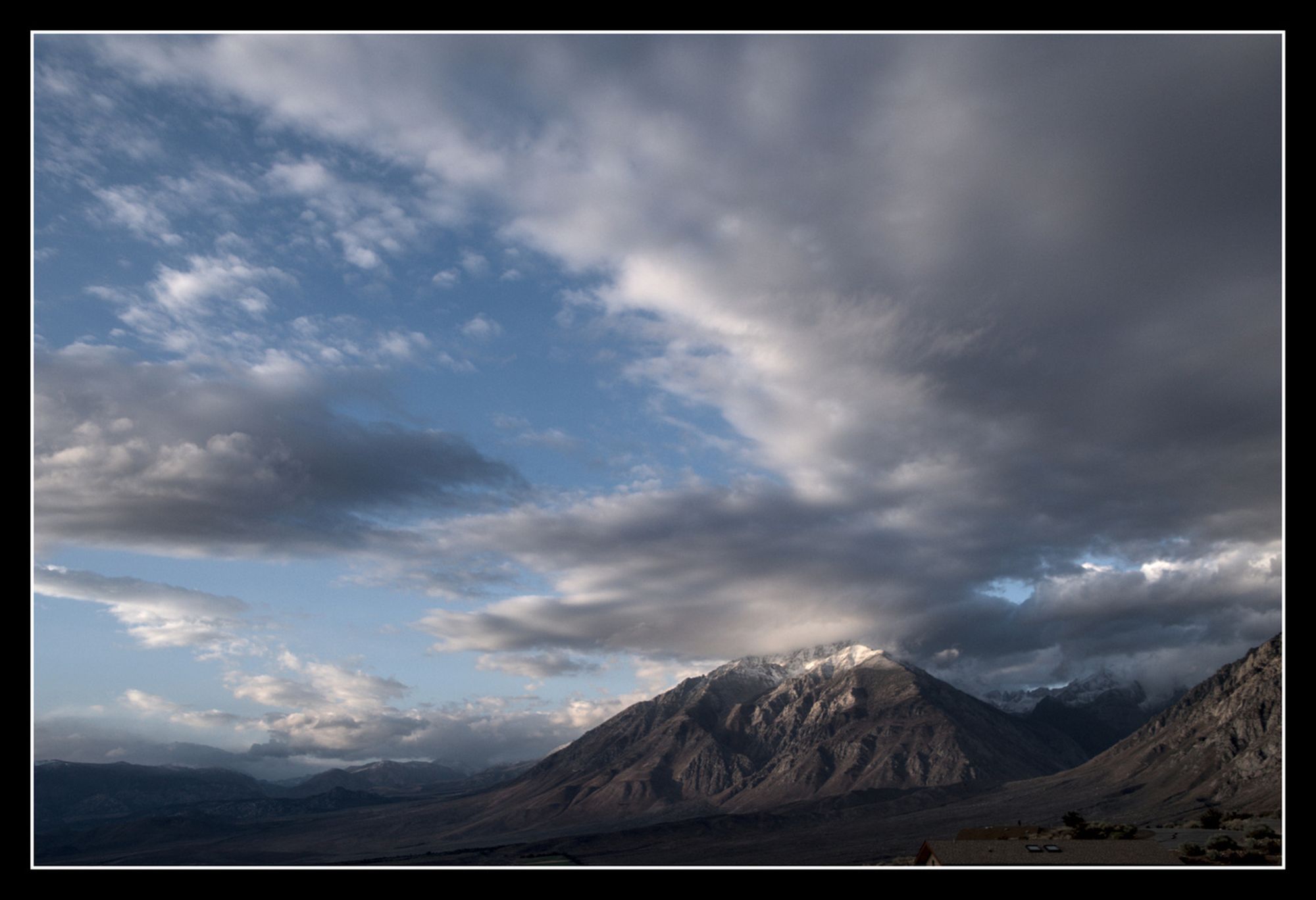 A triangualr mountain with snow on the peak has  a large cloud rising from the top, blue sky to the left, dark cloud to the right.