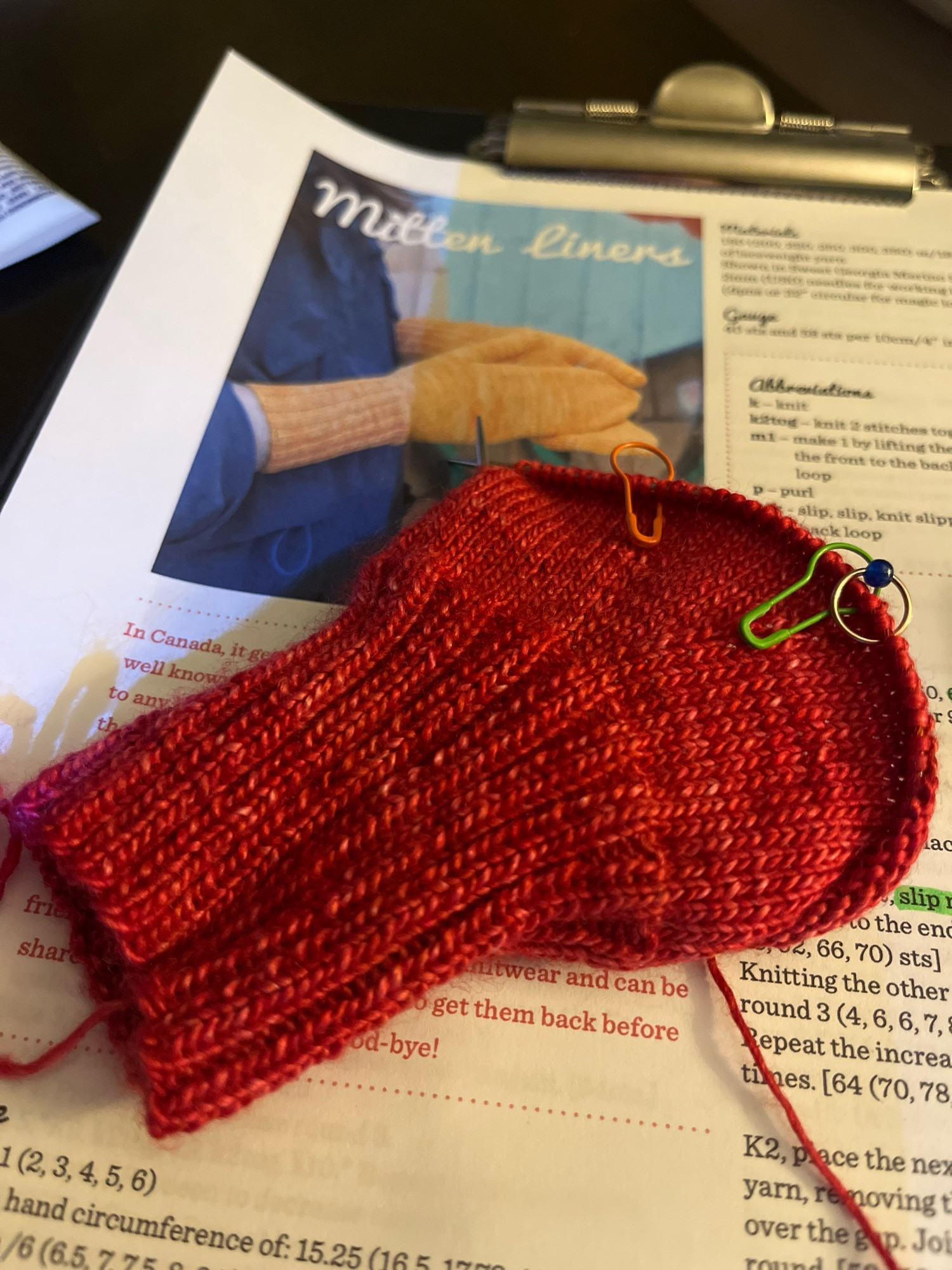 Berry coloured work in progress mittens with several bright stitch markers. They are sitting on top of the pattern.