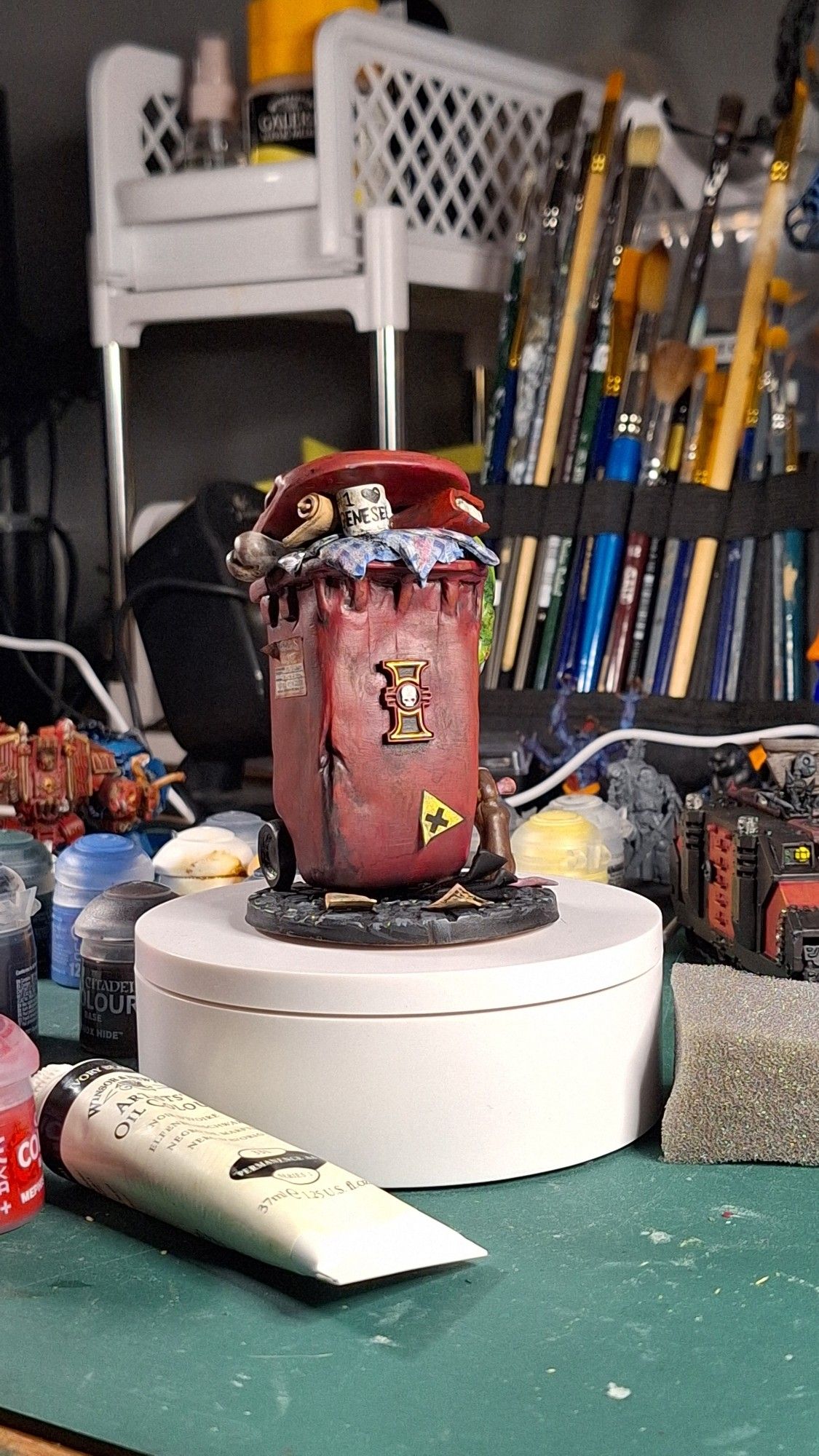 9:16 colour photo. Cluttered. There is a small sculpted wheelie bin on a white turn table. The bin has the Inquisition logo from Warhammer 40k. There are hazard stickers.