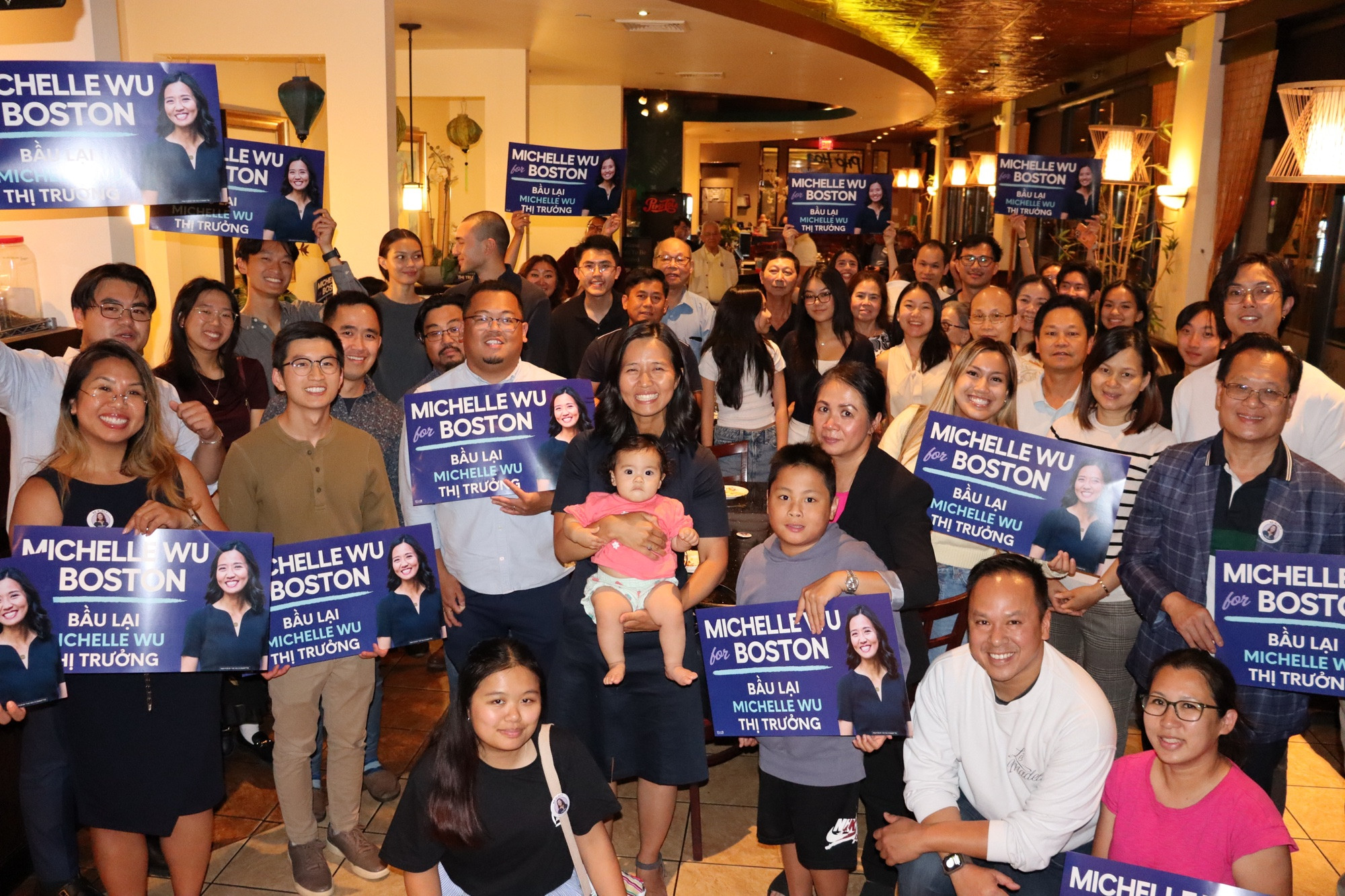 A large crowd of people stand together in Pho Hoa Restaurant holding Michelle Wu for Boston signs with Mayor Wu and Baby Mira in the middle. 