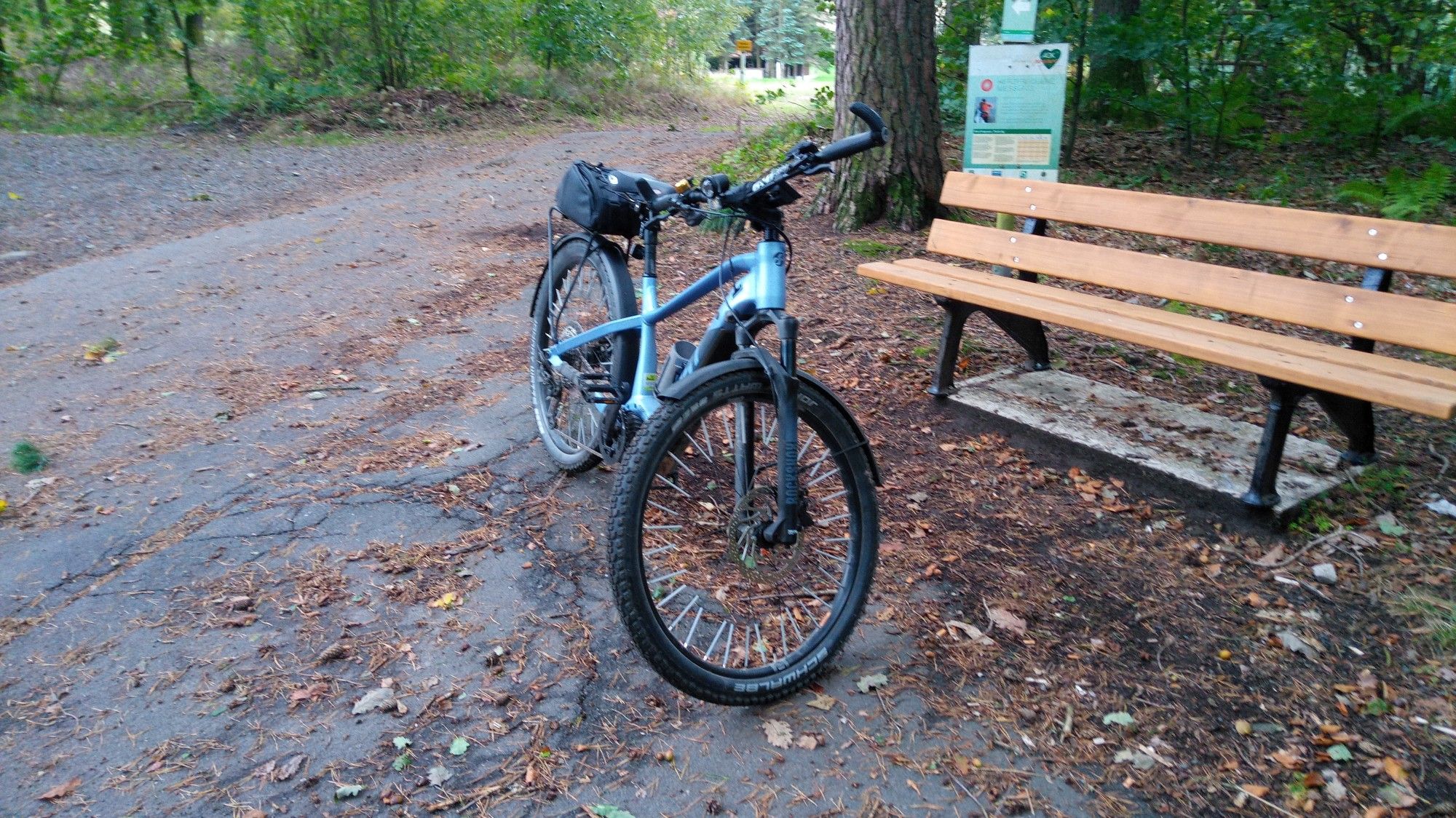 Ein mattblaues SUV Mountainbike vor einer Holzbank im Wald, dahinter sind mehrere Wanderwegweiser.