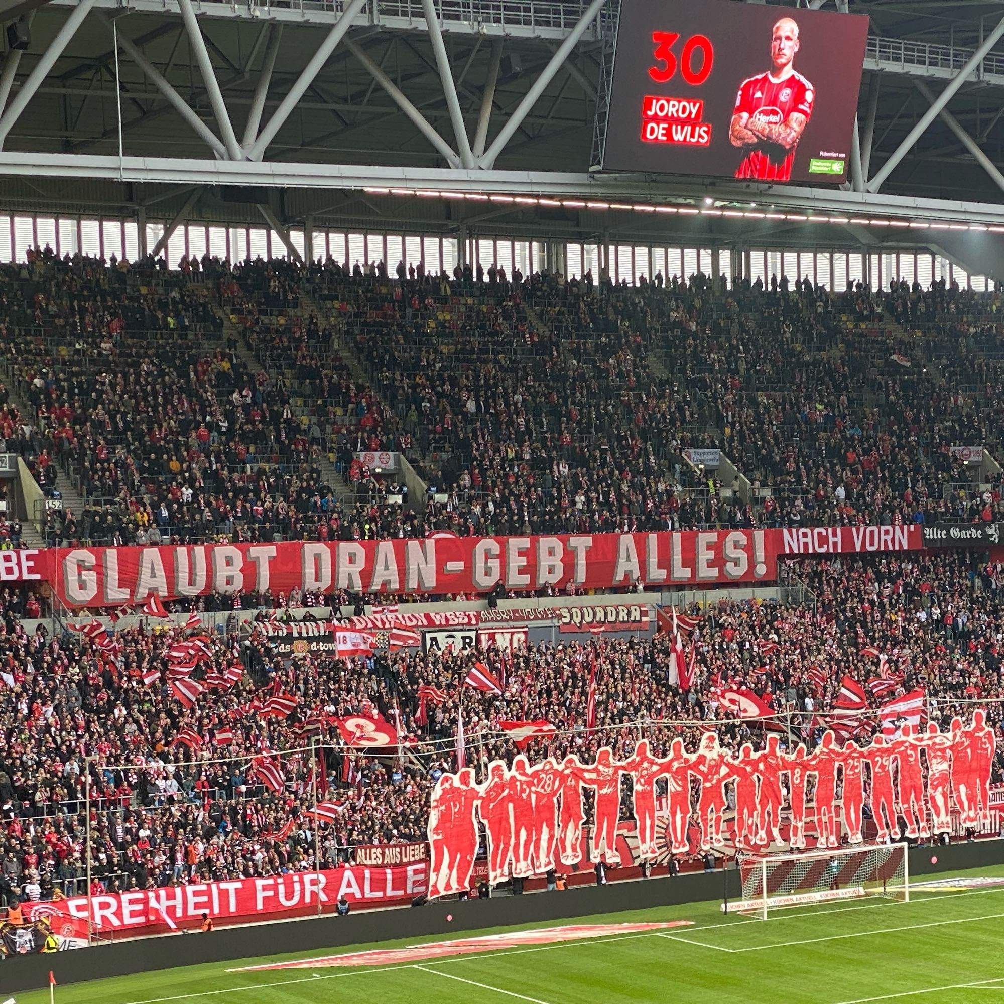 Blick auf den Unterrang der Düsseldorfer Südtribüne.
Choreo: überlebensgroße rote spielersilhouetten, dazu ein Spruchband: glaubt dran- gebt alles