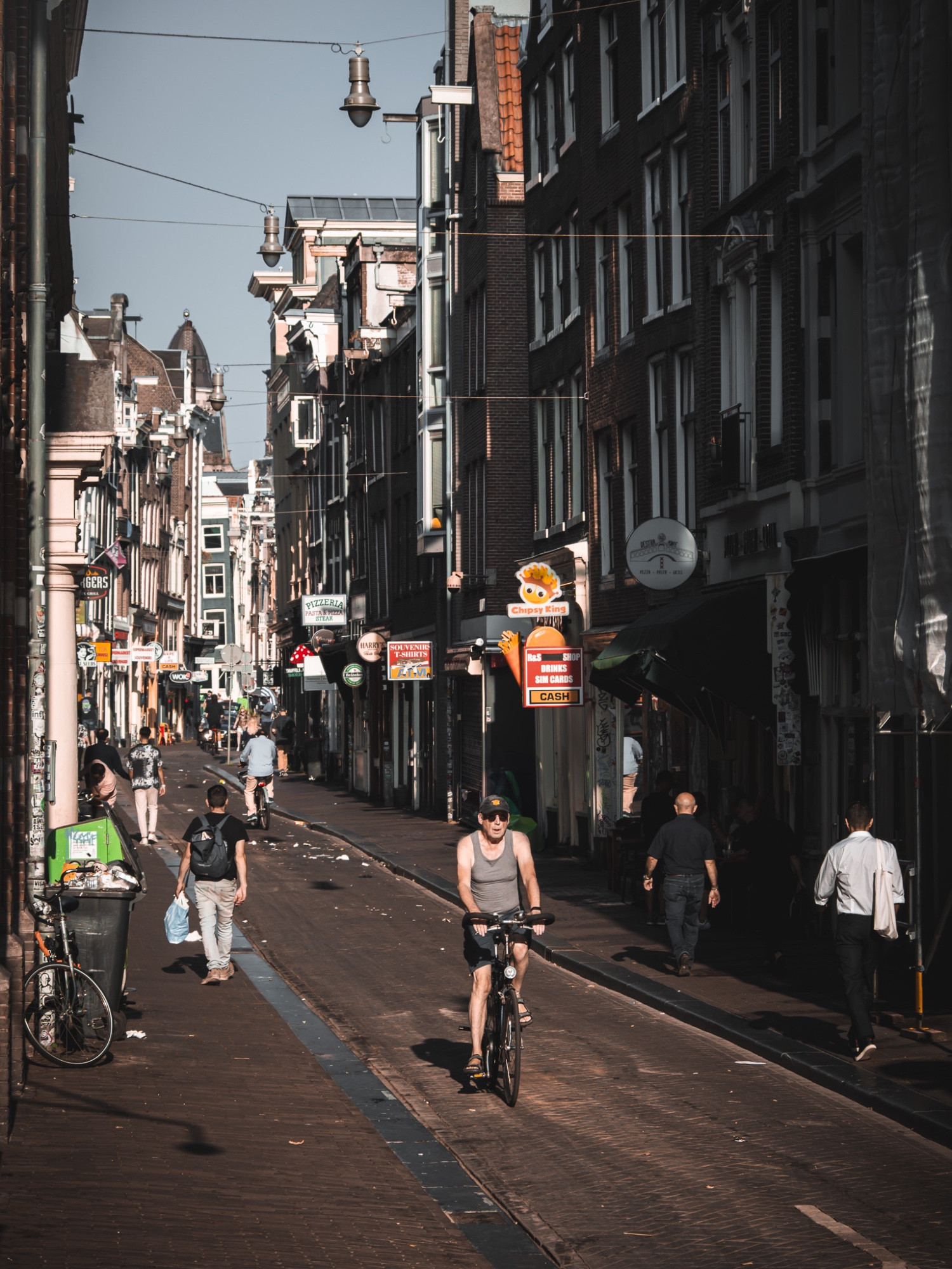 A narrow street in Amsterdam on an early sunny Sunday morning. A man wearing a tank top and sunglasses rides a bicycle down the brick-paved street, while a few pedestrians walk nearby. The street is lined with tall, traditional buildings and various shop signs. The scene is quiet, with the sun casting long shadows.